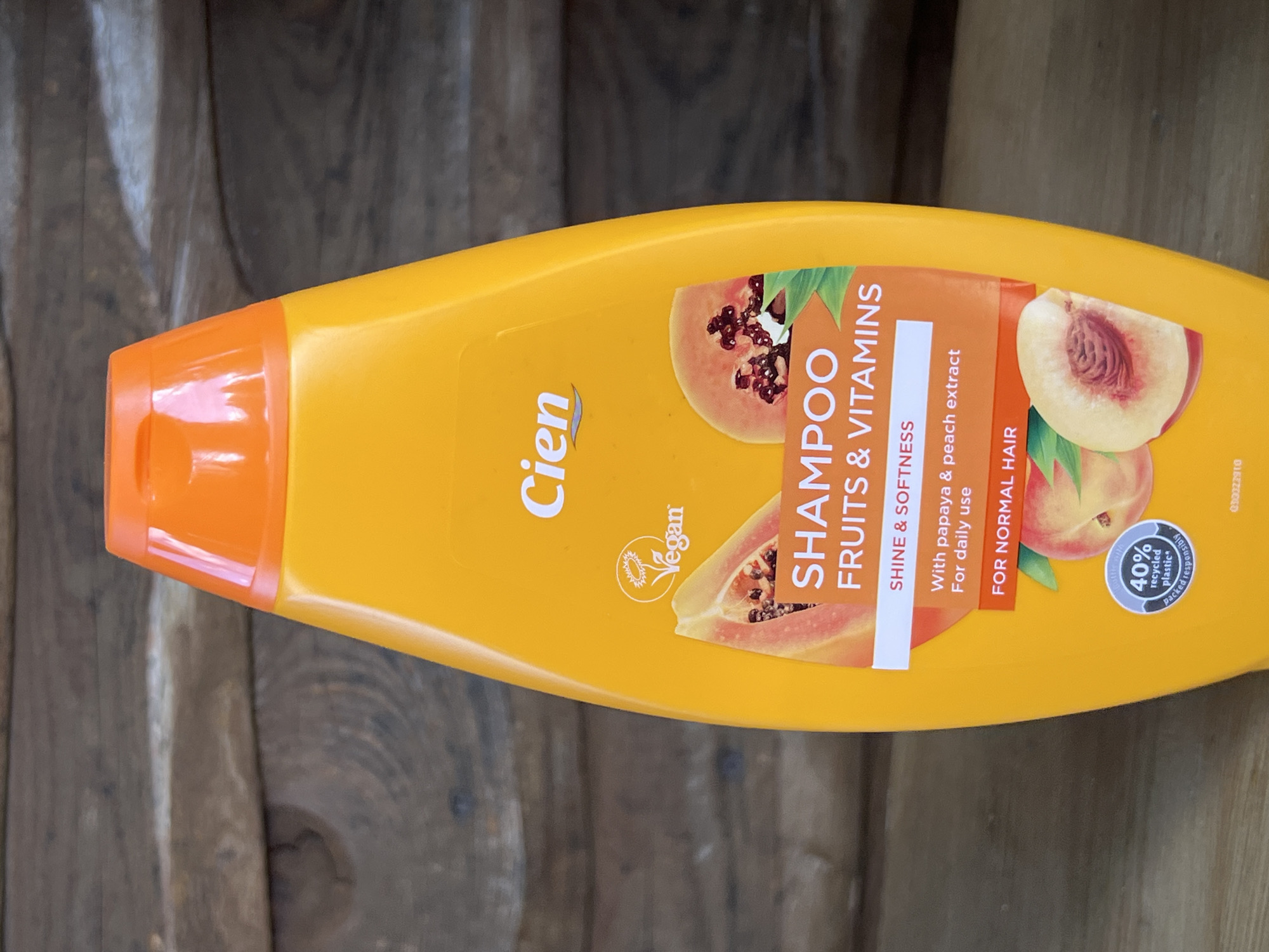 A close-up, eye-level shot shows a bright orange plastic bottle of Cien shampoo, set against a weathered, dark brown wooden plank background. The bottle is positioned centrally in the frame, with its vibrant color contrasting sharply with the rustic texture of the wood. The shampoo bottle has an orange cap and a label featuring stylized graphics of papaya and peach slices, along with text indicating "SHAMPOO FRUITS & VITAMINS," "SHINE & SOFTNESS," and "With papaya & peach extract For daily use For normal hair." A small circular emblem at the bottom of the bottle highlights "40% recycled plastic packed responsibly." The background appears to be an outdoor wooden surface, possibly a fence or wall, with visible grain and slight weathering. The lighting is natural and casts subtle shadows. No people or animals are present in the image. The scene is static and appears to be a product shot. The location in Daulėnai, Lithuania is not discernible from the image itself.