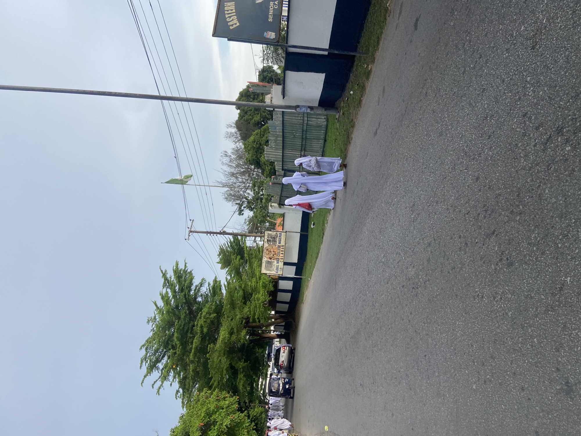 The image shows a street scene in Calabar, Nigeria. A group of people in white robes are walking on the right side of the road, near the Naval War College of Nigeria. A sign for "Eastern High School" is also visible. A green Nigerian flag is flying in the background. A row of parked cars lines the left side of the road, under a canopy of trees. Overhead power lines run across the sky. The road is paved with asphalt. The sky is overcast.