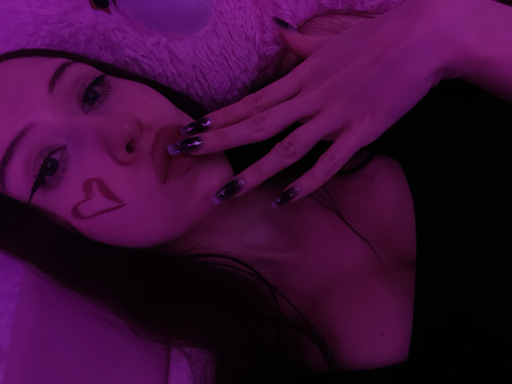 The image is a selfie of a young woman in Votkinsk, Russia. She has long, dark hair and is wearing a black top. A red heart is drawn on her left cheek. Her nails are long and painted dark with a subtle shimmer. She is holding her right hand to her mouth. The scene is lit with purple light, creating a moody atmosphere. A fluffy pink object, possibly a blanket or stuffed animal, is visible in the background.