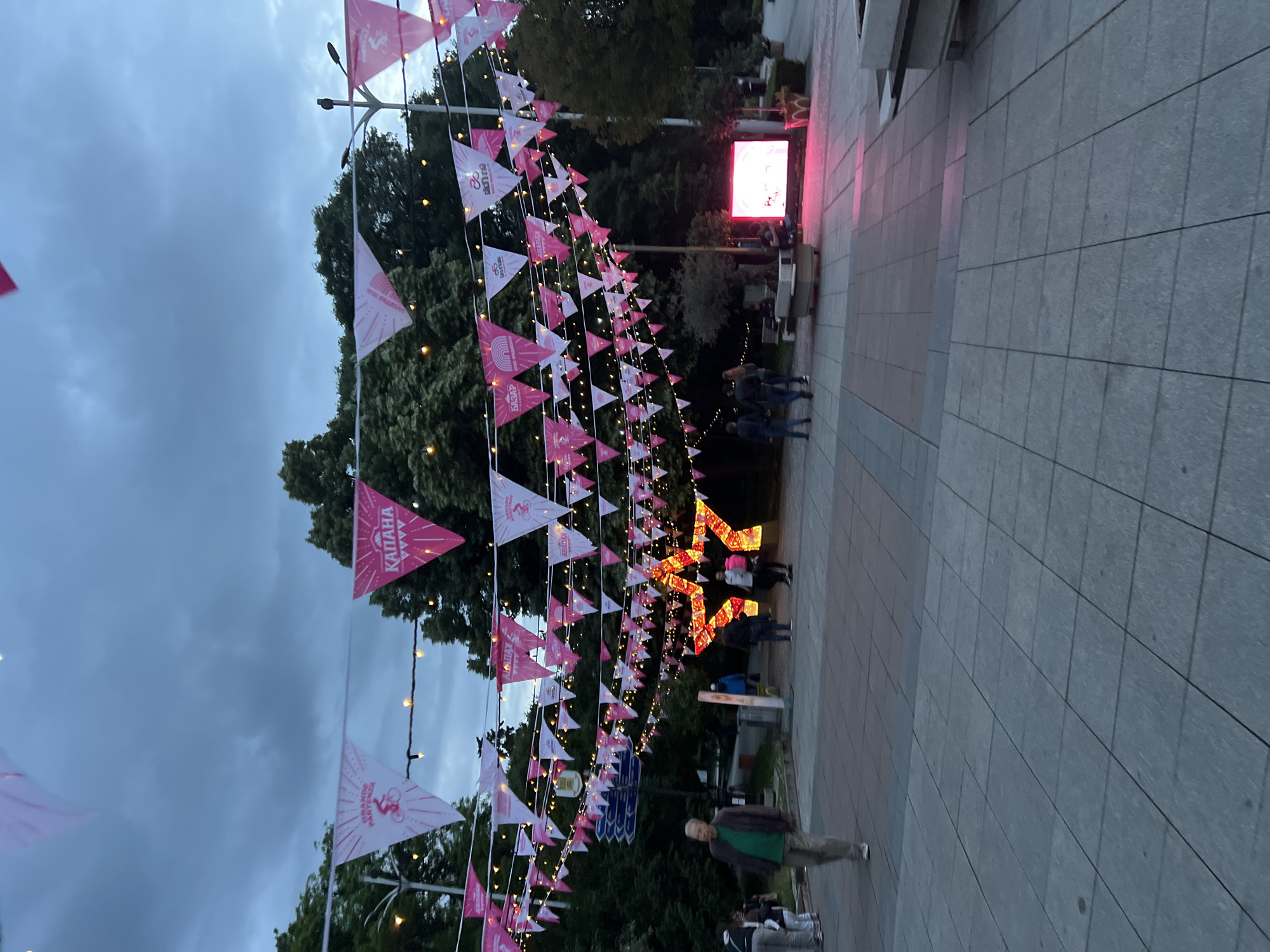 The image depicts a street scene in Plovdiv, Bulgaria. Bunting, with triangular pink and white flags, stretches across a pedestrianized walkway. Some flags feature the word "КАПАНА". String lights are interspersed with the bunting. In the background, a video screen glows with a red display. The ground is paved with large square tiles. Several people are walking or standing in groups along the walkway. One man is in the foreground, wearing a green shirt and tan pants. Trees are visible in the background. The sky is overcast and dusky.