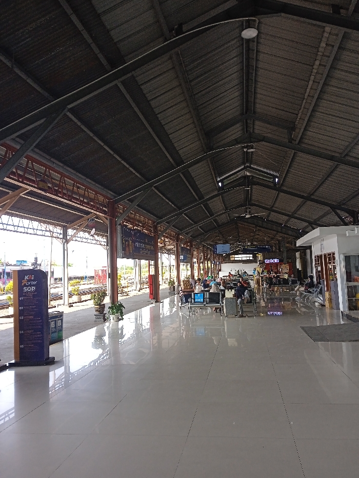 The image depicts the interior of a train station in Cangkring, Indonesia, characterized by a spacious, open layout. A high, vaulted roof constructed from corrugated metal panels supported by dark metal trusses and pillars covers the area. The floor is composed of large, glossy light-colored tiles, reflecting the overhead structure and ambient light.
On the left side of the frame, a train platform is visible with tracks beyond. A dark blue vertical sign labeled "KAI Porter SOP" stands near the platform edge, adjacent to two trash receptacles. Several potted plants are positioned along the platform. In the mid-ground, a waiting area features multiple dark-colored benches arranged in rows, occupied by approximately 15 to 20 individuals. These passengers are primarily seated, some attending to personal devices, others conversing, and some with visible luggage such as suitcases.
Towards the right side, a small white building, possibly an office or kiosk, with brown-framed windows is present. Overhead, numerous circular and linear light fixtures are mounted on the roof structure, along with at least two visible ceiling fans. Digital display screens are observable further down the waiting area. Natural light enters from the open sides of the station structure.