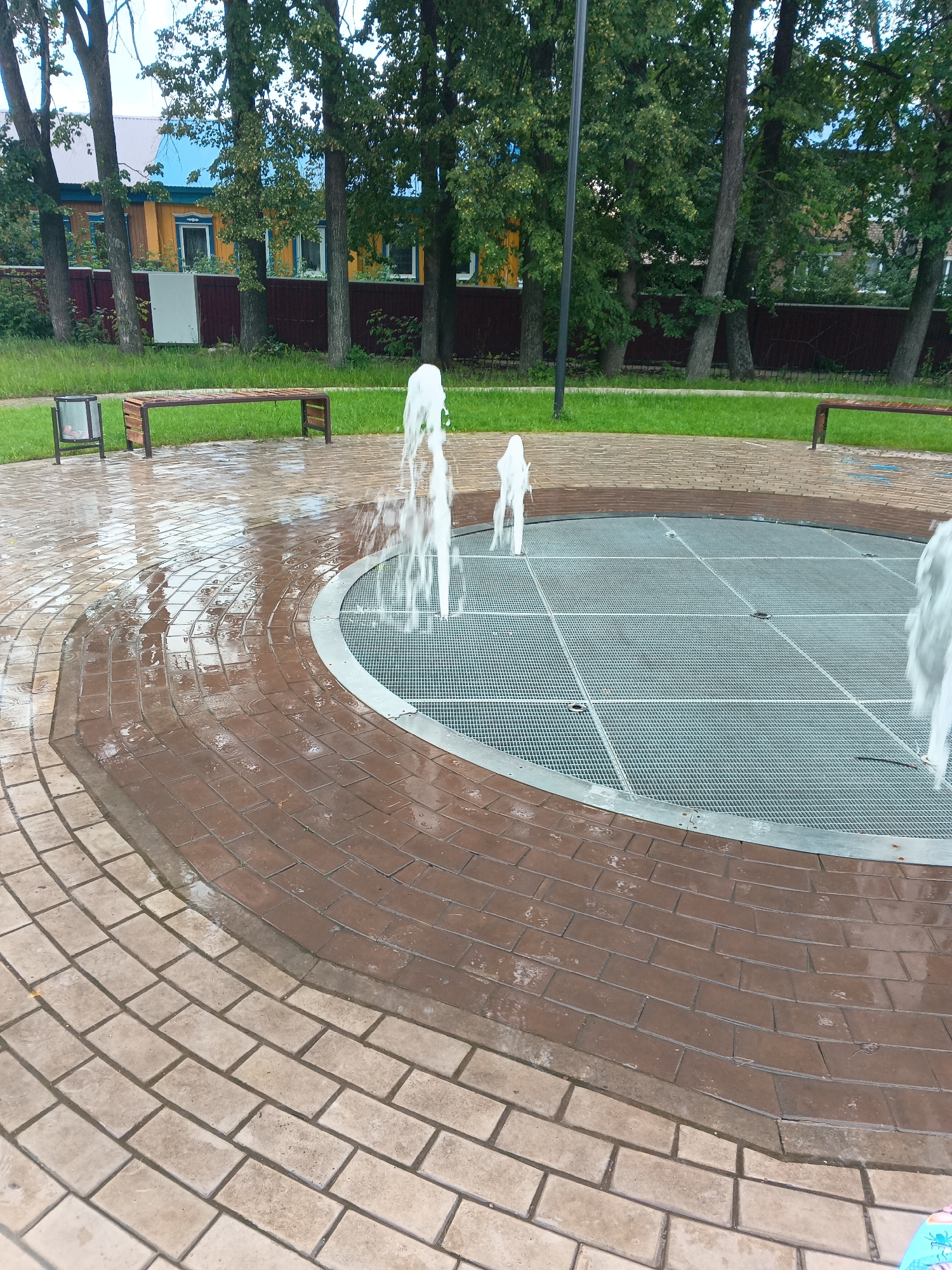 The image captures an outdoor public space in the city of Tolbazy, Russia, featuring a circular dry fountain. The fountain's surface is a metal grate with a grid pattern, surrounded by brick paving. Water jets are actively spraying upwards from the grate, creating a dynamic visual element. The paving stones are wet, indicating recent activity or rainfall. In the background, several trees with lush green foliage are visible, with a wooden fence and a multi-colored residential building behind them. A park bench and a trash receptacle are situated on the grass beyond the paved area, suggesting a recreational setting. The overall scene is one of a well-maintained urban park or public square.