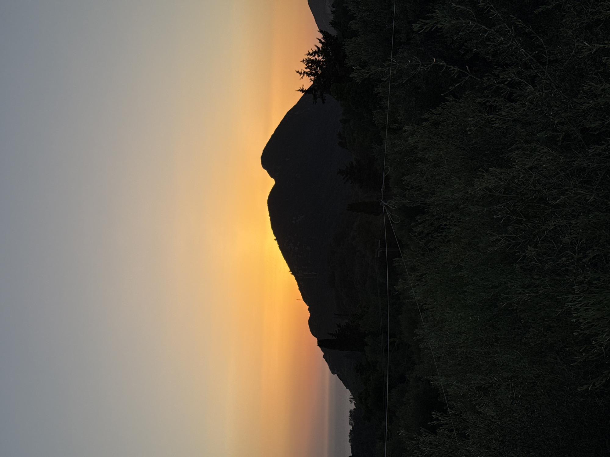 A tranquil sunset scene unfolds over a mountainous landscape with the sea visible in the distance. The sky transitions from a soft grey at the top to a vibrant orange and yellow gradient as it meets the silhouetted mountain range. The mountain peaks are dark and imposing, with a peak in the center where the sun is setting, casting a bright glow behind it. Dense, dark foliage, likely trees and bushes, covers the lower slopes of the mountains and stretches across the foreground, obscuring much of the ground. A few thin, dark wires or ropes are visible stretched horizontally across the lower part of the image, suggesting some form of man-made structure. The overall impression is one of natural beauty and peaceful seclusion. This scene appears to be located in Kokkini, Greece.