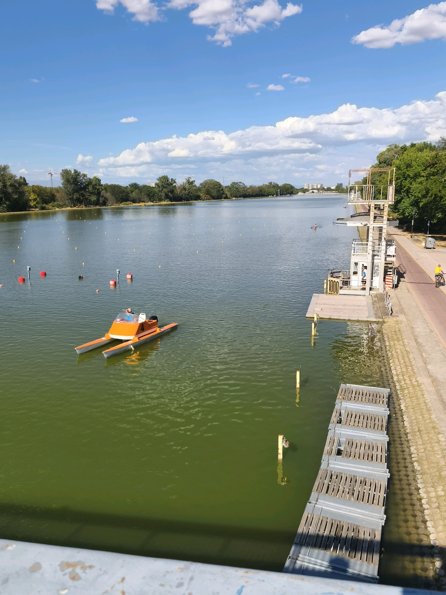 The image depicts a wide, calm body of water with a clear blue sky filled with fluffy white clouds. On the left side of the water, a small orange and white catamaran-like boat with a person at the helm is visible, alongside several red and white buoys. The right side of the water features a concrete embankment with a paved walkway and a tall, multi-tiered structure resembling a diving or observation platform with stairs and railings. A person on a bicycle is seen on the walkway, and a yellow trash bin is positioned near the embankment. In the distance, a treeline borders the water, with buildings and what appears to be a crane visible beyond the trees. A long, wooden boardwalk or pier extends from the embankment into the water on the right side of the frame. The overall scene suggests an outdoor recreational area, possibly a park or a sports facility located on a lake or large pond.