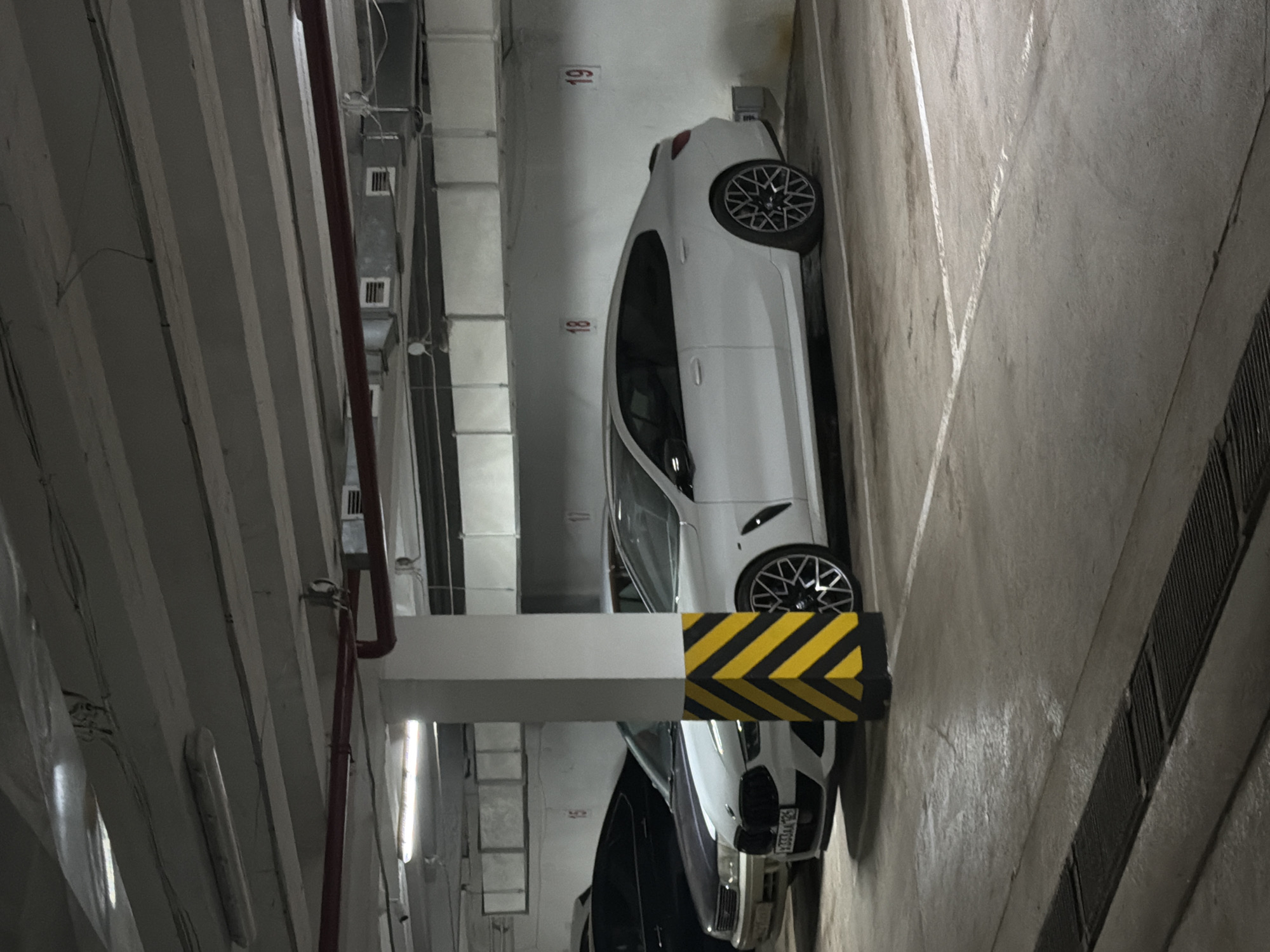 The image shows a parking garage in the city of Krasnoyarsk, Russia. Two cars are parked in adjacent spaces. One car is white and the other is dark. Both cars are BMWs. The parking spaces are marked with numbers. The number "19" is visible near the white car. A structural column with yellow and black hazard markings is positioned between the two cars. The garage environment is dimly lit and appears to be constructed of concrete. The license plate of the dark colored car is visible.