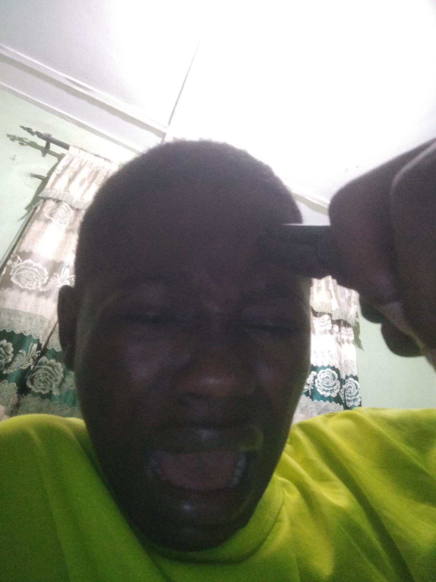 A close-up photograph depicts a dark-skinned person with a shaved head wearing a bright green t-shirt. They are grimacing with their eyes closed and mouth open. A dark object, possibly a pen or marker, is held to their forehead. In the background, there is a patterned curtain. No specific details indicate the location to be Shangisha, Nigeria.