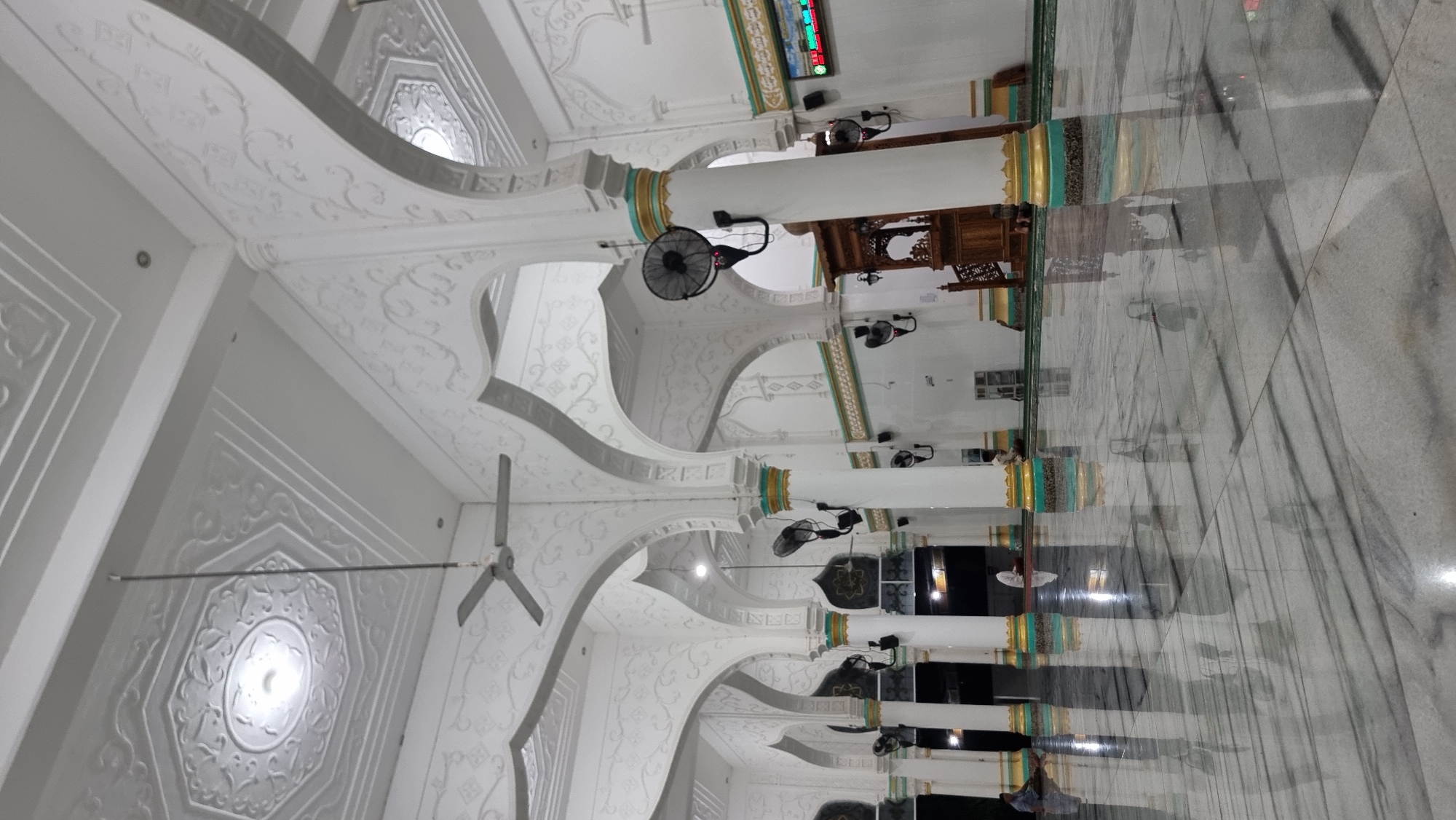 The image shows the interior of a mosque in Lapang Timu, Indonesia. The mosque features white vaulted ceilings with intricate embossed patterns and a large octagonal recessed panel with a central light fixture. White cylindrical columns with gold and teal decorative bands support the arches. Several black wall-mounted fans are visible throughout the space. The floor is made of polished marble with a grey and white veined pattern, reflecting the ceiling and architectural elements.
In the foreground, the reflective floor dominates the view. In the middle ground, several people are seen sitting or kneeling on prayer mats. One individual is dressed in white and appears to be praying near a dark doorway. Another person in a light-colored garment is sitting on the floor to the left of the frame.
To the right, a wooden pulpit, or *mimbar*, with intricate carvings is positioned against a white wall. Above the pulpit, a digital display board shows colored text. The background shows more arches and columns, creating a sense of depth within the mosque. The overall lighting is bright, suggesting interior illumination.