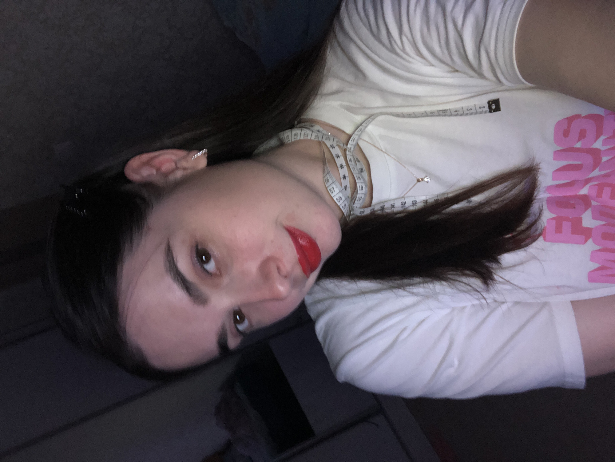 A woman is posing for a picture. She has fair skin, dark hair, and red lipstick. She's wearing a white t-shirt with pink lettering and a tape measure around her neck. The environment appears to be dimly lit, possibly indoors. The image location is unknown and not related to Noviy, Russia.
