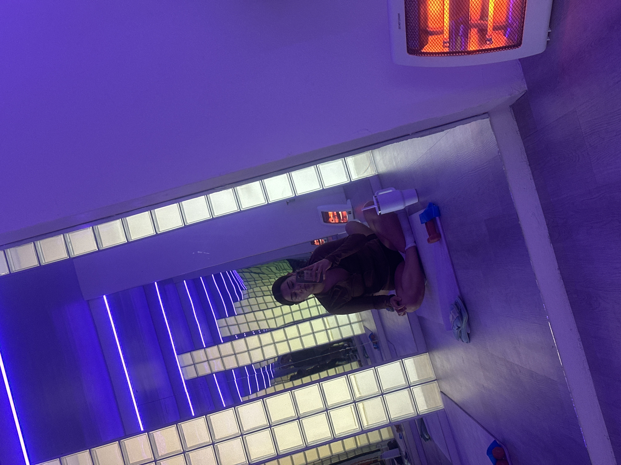 A person is taking a selfie in a bathroom with infinity mirrors in San Bernardino Tlaxcalancingo, Mexico. The room is illuminated with blue and white neon lights. The individual is wearing a brown top and has a dark beanie. There is a radiator and a hair dryer on the wall. Blue cleaning supplies are visible near the floor.