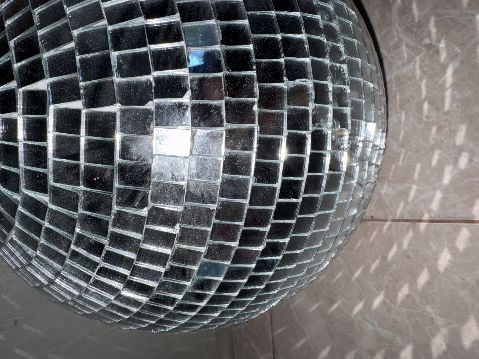 The image prominently features a large disco ball, covered in an array of small, square, reflective mirror tiles. The ball occupies the left and central portions of the frame, showcasing its distinct curved surface. Many of the individual mirror tiles exhibit varying degrees of reflectivity, with some appearing dark due to shadows or reflections of darker areas, while others gleam brightly from direct light exposure. Visible grout lines and some wear or dust are present between the tiles.
To the right of the disco ball, a light-colored, possibly tiled or textured wall or surface is visible. This surface is illuminated with numerous scattered, square-shaped reflections of light emanating from the disco ball, indicating a direct light source hitting the ball off-frame. The scene is static, capturing the aesthetic qualities of the disco ball and its light-reflecting properties. No people or specific activities are depicted. The setting appears to be indoors. There is no visible text in the image. The location context for this scene is North Kuta, Indonesia.