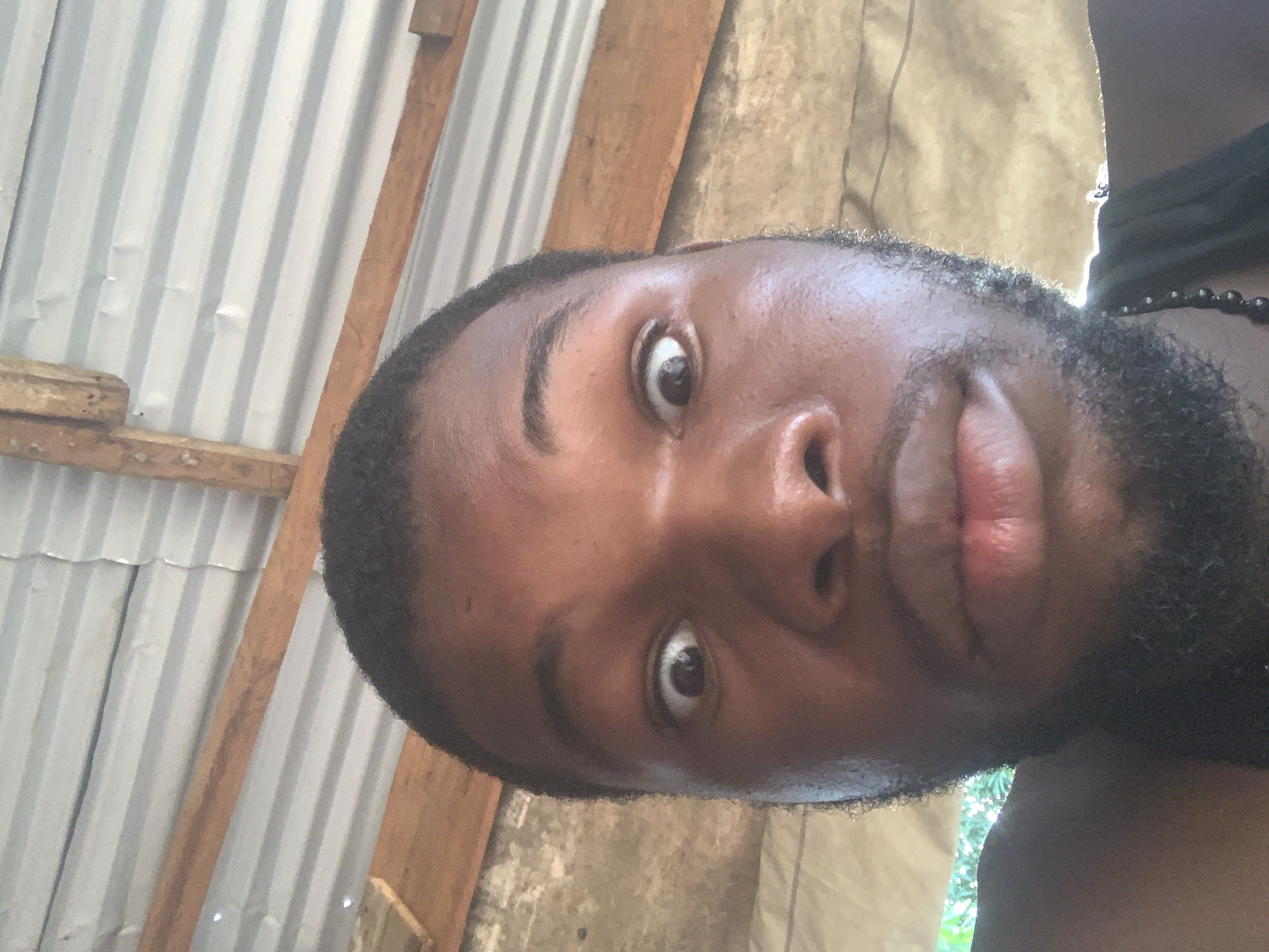 A close-up selfie features a dark-skinned man from the shoulders up, captured at a tilted angle. He looks directly at the camera with wide, round eyes, conveying a surprised or startled expression. His lips are slightly pursed. He has short, dark hair and a short, dark beard. A dark beaded necklace is visible around his neck, and part of a dark garment is seen on his right shoulder.
The background depicts a rustic indoor or semi-sheltered environment. It is dominated by vertical, light-colored corrugated metal sheets, likely forming a wall or ceiling, which show minor dents. Rough-hewn wooden beams or planks are also visible, providing structural support to the corrugated material. A small wooden block rests on one of these beams in the lower left background. The scene is illuminated by natural light, casting highlights on the man's skin. There is no visible text in the image.