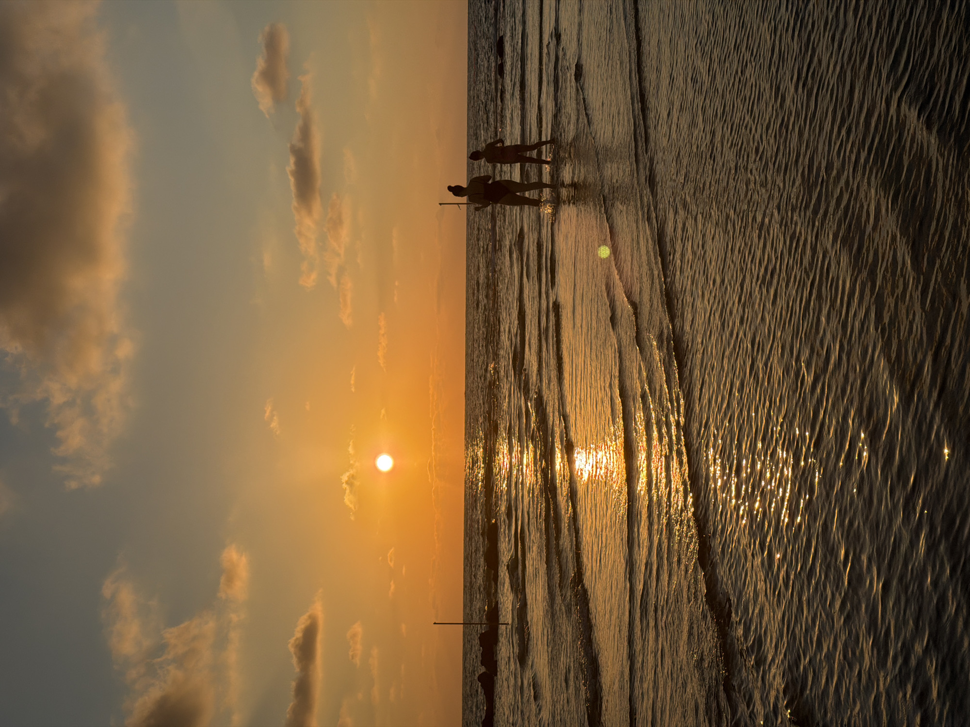 A silhouette of two figures, appearing to be an adult and a child, stand in the shallow water of the Mediterranean Sea at sunset. The adult is wearing what appears to be a swimsuit. They are facing towards the setting sun, which casts a warm, golden glow across the water and the sky. The sky is filled with scattered clouds, tinged with orange and yellow. Gentle waves lap at the shore, creating ripples that reflect the sunlight. In the distance, a series of dark, irregular shapes, possibly rocks or a breakwater, are visible near the horizon. A tall, thin pole emerges from the water to the left of the figures. The scene suggests a peaceful moment at the end of the day, likely on one of Bat Yam's beaches.