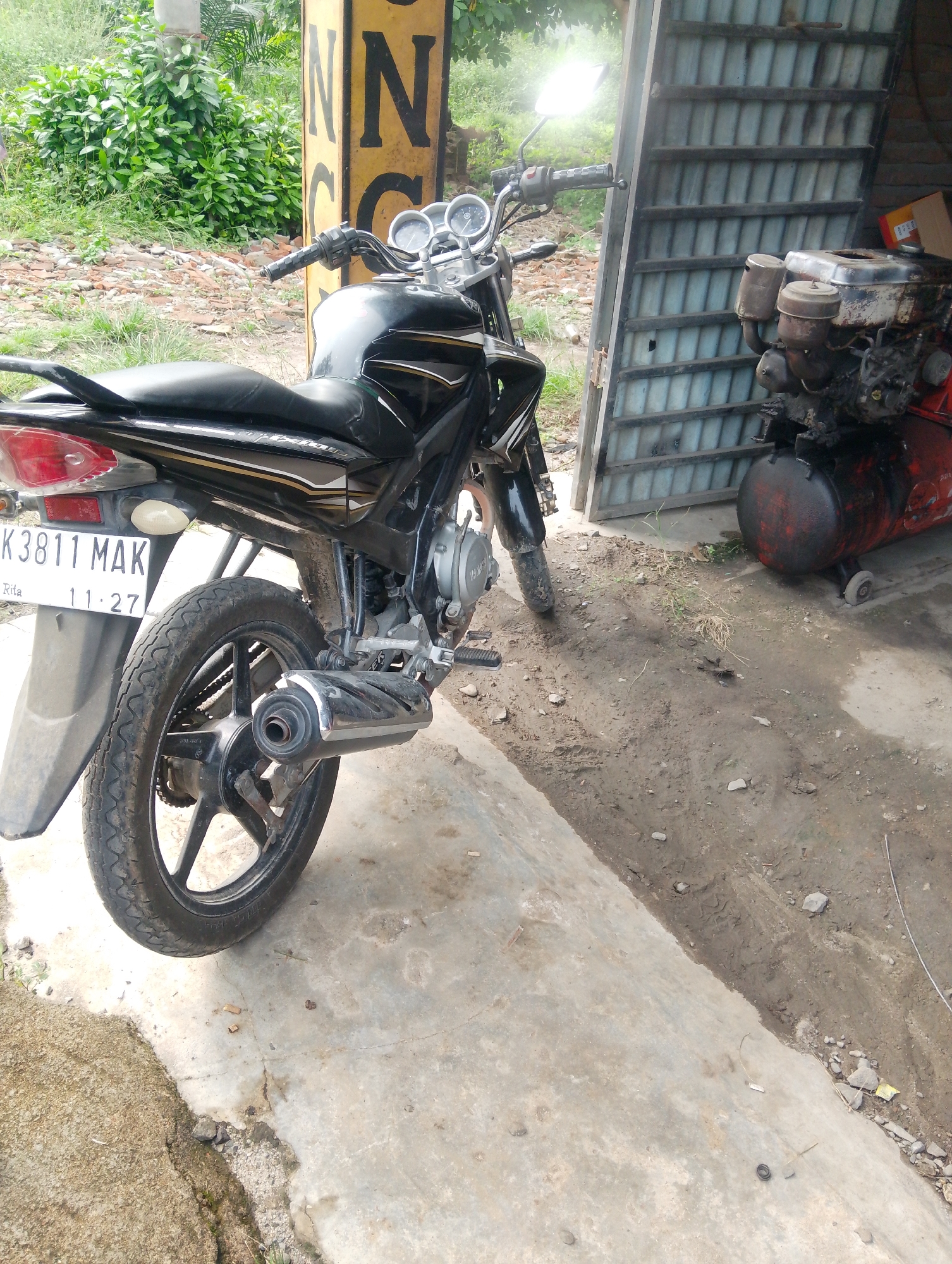 A black motorcycle is parked next to a corrugated metal structure in what appears to be an outdoor workshop or storage area. The motorcycle is viewed from the rear and right side, with its license plate visible, reading "BK 3811 MAK" and "11.27". To the right of the motorcycle, a red and black engine with two exhaust pipes and air filters is situated. The ground is a mixture of concrete and dirt, with some scattered debris. In the background, lush green foliage can be seen, suggesting an outdoor location. The overall scene appears to be a simple, functional workspace.