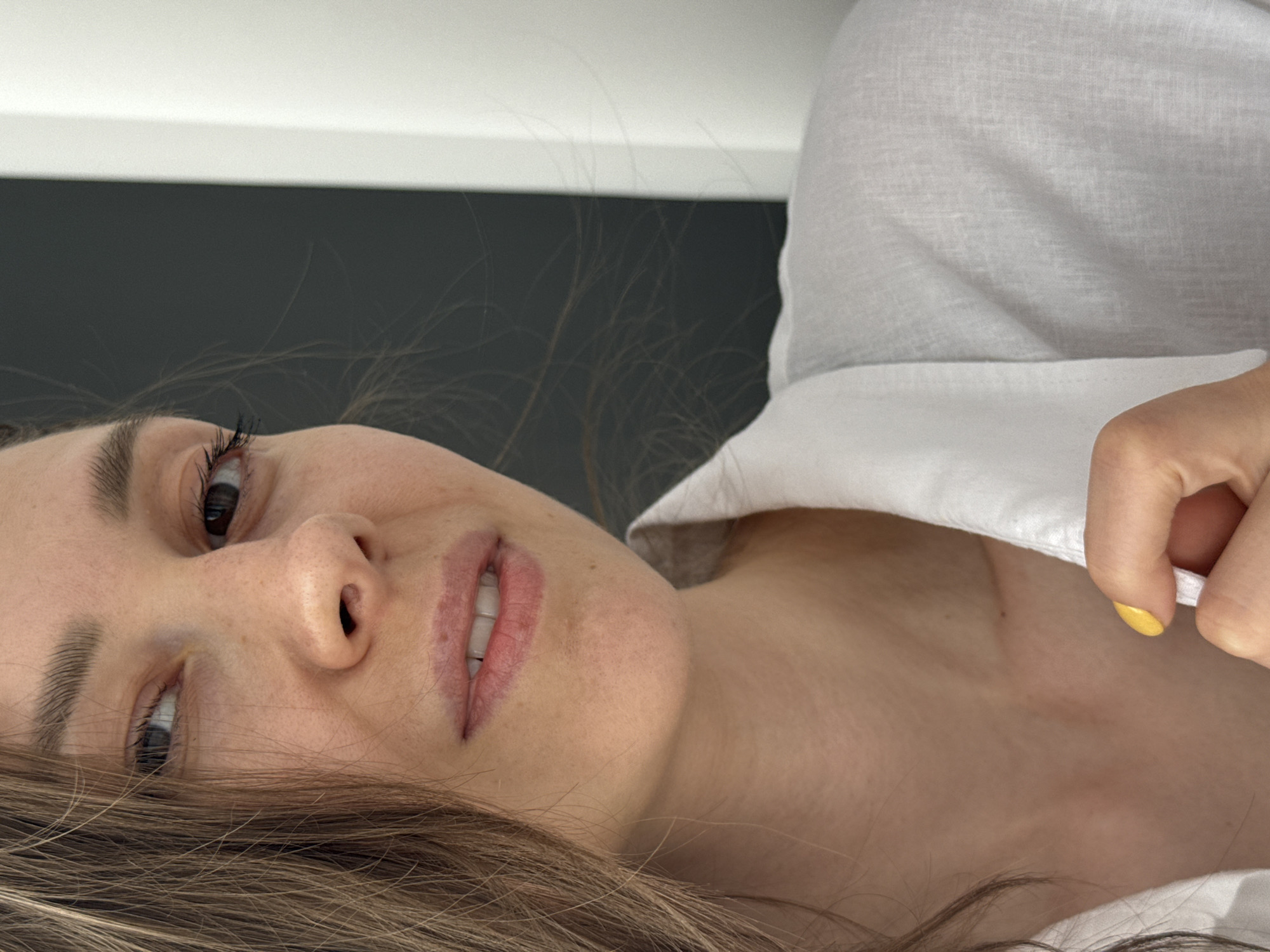 A close-up, tilted shot depicts a woman with fair skin, brown hair, and brown eyes. She is wearing a white shirt and has a yellow nail polish on one of her fingers. The background appears to be a dark grey or black wall with a white shelf or surface above it. There's no information to confirm a location in Chaikovsky, Russia.