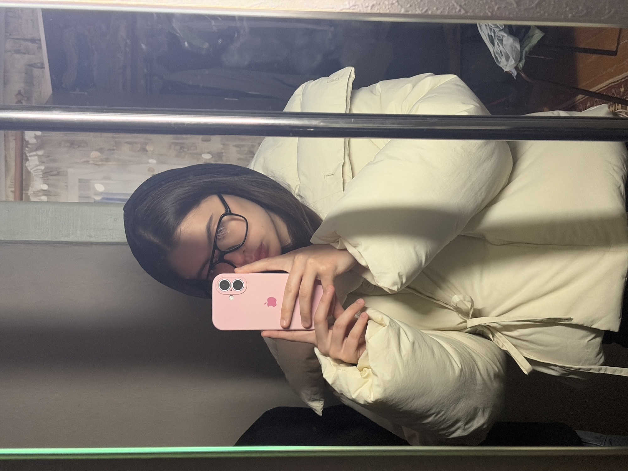 The image captures a mirror selfie taken indoors, rotated 90 degrees clockwise. The main subject is a young individual, likely female, with dark hair, wearing dark-rimmed glasses, and a light cream or off-white puffy jacket or coat. They are holding a pink iPhone with the prominent Apple logo and dual camera lenses visible on the back, suggesting an iPhone 11 or 12 model. The person is looking towards the phone or just past it, their face partially obscured by the device.
A polished metallic horizontal bar is visible in the foreground, extending across the mid-section of the image, reflecting light. Through the mirror's reflection, the background reveals an indoor setting, possibly a bedroom or living space in Chaikovsky, Russia. Elements visible in the reflection include what appears to be a bed with light-colored pillows or duvet, and a wall with faded or textured wallpaper. In the upper right of the reflection, some folded clothes (grey and green fabric) are stacked, and a glimpse of a patterned rug or carpet can be seen. The overall lighting is somewhat dim, creating a subdued atmosphere indoors. No specific text is visible other than the Apple logo on the phone.