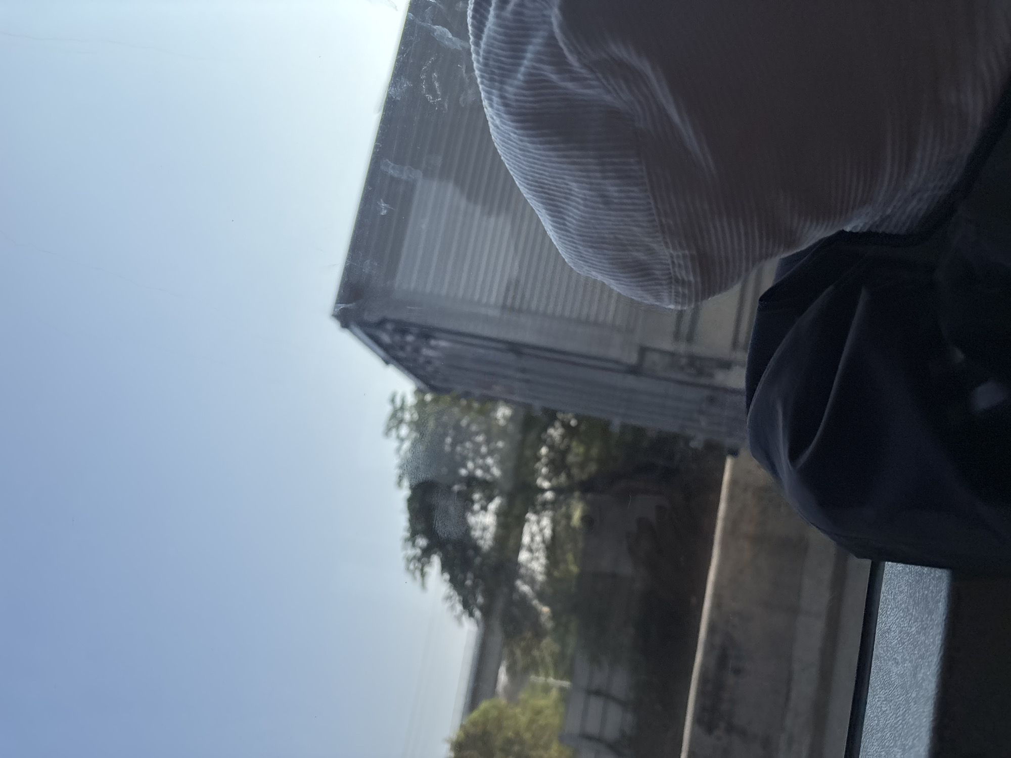 The image appears to be taken from inside a vehicle, looking out through a window. The foreground shows the back of a person's head, wearing a light-colored corduroy cap, and a dark jacket. The background is blurred, but visible elements include a large trailer truck, trees, and what appears to be a highway overpass or bridge structure. The sky is a clear, light blue. The overall impression is that of a journey, possibly on a road trip, with the landscape passing by. Without further context or identifying markers, it is not possible to determine the specific location within Jayamukti, Indonesia.