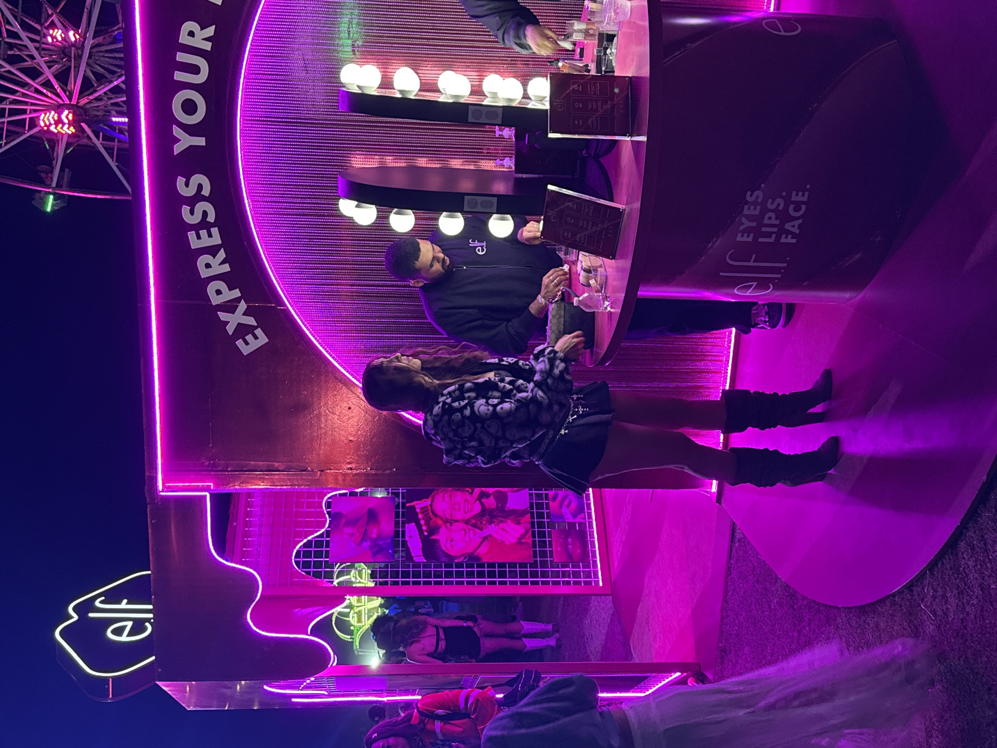 The image depicts a vibrant scene at an e.l.f. Cosmetics event, likely in Thermal, California, based on the surrounding landscape and fairground structures. A tall ferris wheel is partially visible in the background, illuminated with colorful lights against a dark sky. In the foreground, there's an e.l.f. branded booth with neon signs and bright pink and purple lighting. A woman is standing at the counter inside the booth interacting with a man behind the counter who wears an e.l.f. branded jacket. The counter is equipped with lighted makeup mirrors and promotional materials. On the side of the counter it reads "elf EYES. LIPS. FACE.". The area is crowded with other people visible inside the booth. The event space seems to be set up outdoors, likely during an evening.