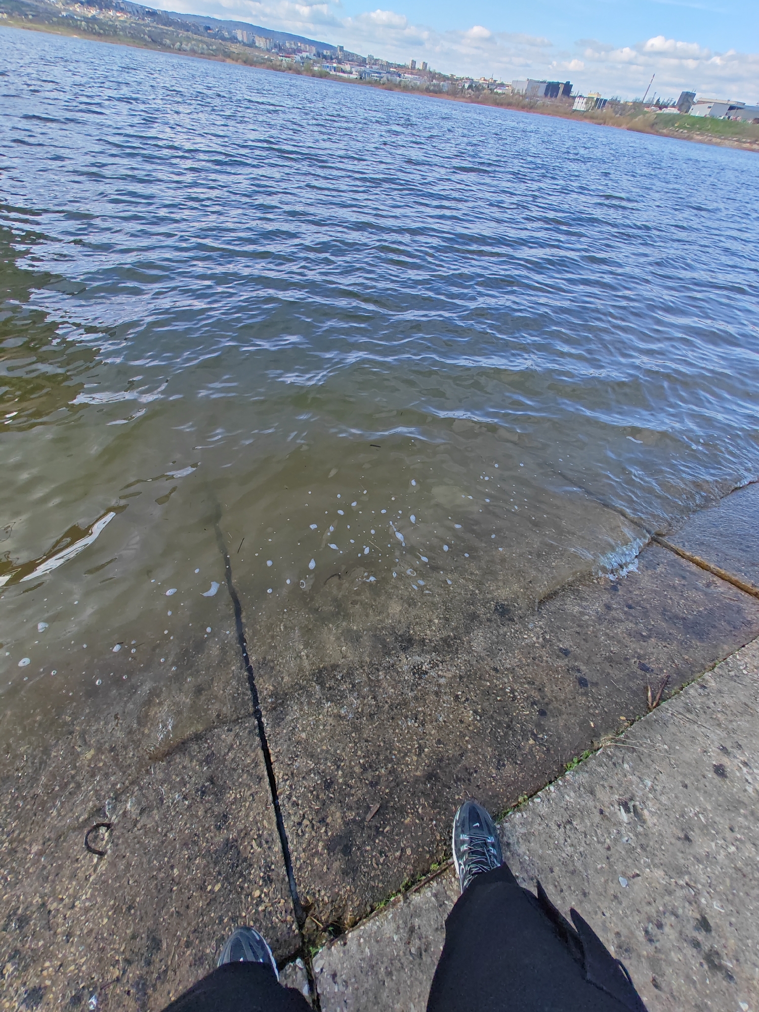 The image captures a person standing on a concrete embankment at the edge of a body of water, possibly a lake or wide river, on a sunny day with a blue sky and some clouds. The water is lapping against the concrete edge, with some submerged areas. The person is wearing black trousers and dark-colored sneakers with light-colored laces. The background shows a cityscape with buildings and hills, suggesting an urban location.
The image was most likely taken in the city of Shumen, Bulgaria. The body of water is likely Lake Kyosha.