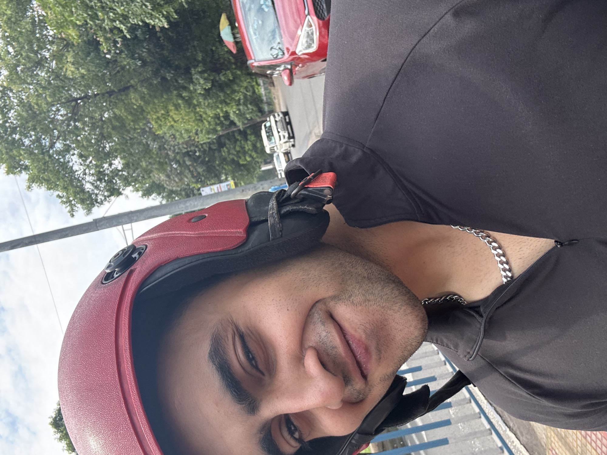 A close-up, ground-level portrait captures a man wearing a red helmet and a dark grey, zip-up top. A silver chain is visible around his neck. In the background, there are green trees, a pole, a red car, and white vehicles. The photo was taken in the city of Kolkata, India.