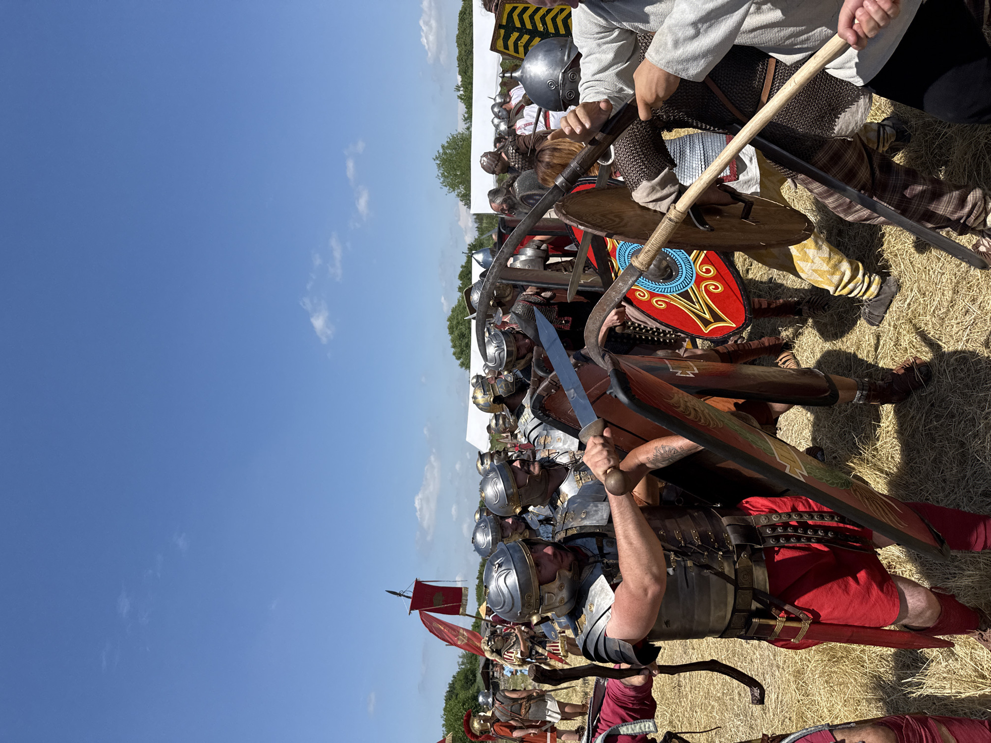 An outdoor scene captures a historical battle reenactment in Gigen, Bulgaria, under a clear blue sky sparsely dotted with white clouds. The ground is covered with dry, light-brown straw or hay. In the immediate foreground, two individuals are engaged in simulated combat. On the left, a person dressed in Roman legionary attire, including a silver segmented metal cuirass (lorica segmentata), a red tunic, red leg wraps, and a helmet, holds a gladius (short sword) with the blade angled towards the right. A large, ornate rectangular shield (scutum) is partially visible to their left. On the right, a person wearing a light-colored tunic, possibly chainmail undergarments, and dark trousers is seen from the torso down, holding a long wooden staff with both hands in a defensive or parrying posture.
In the midground, multiple individuals in full Roman legionary costume, featuring silver helmets, segmented armor, and red tunics, are arrayed. One legionary on the left holds a round, brightly colored shield with a blue and yellow swirling pattern on a red background. Further right, other reenactors wear various historical armors and helmets, some holding distinct weapons like a large, curved bladed polearm (falx) and a shield with yellow and black chevron patterns. A red banner displaying a golden eagle (aquila) on a pole is visible in the background on the far left. White tents are discernible in the distant background, flanked by green trees and foliage, indicating an event setup.
