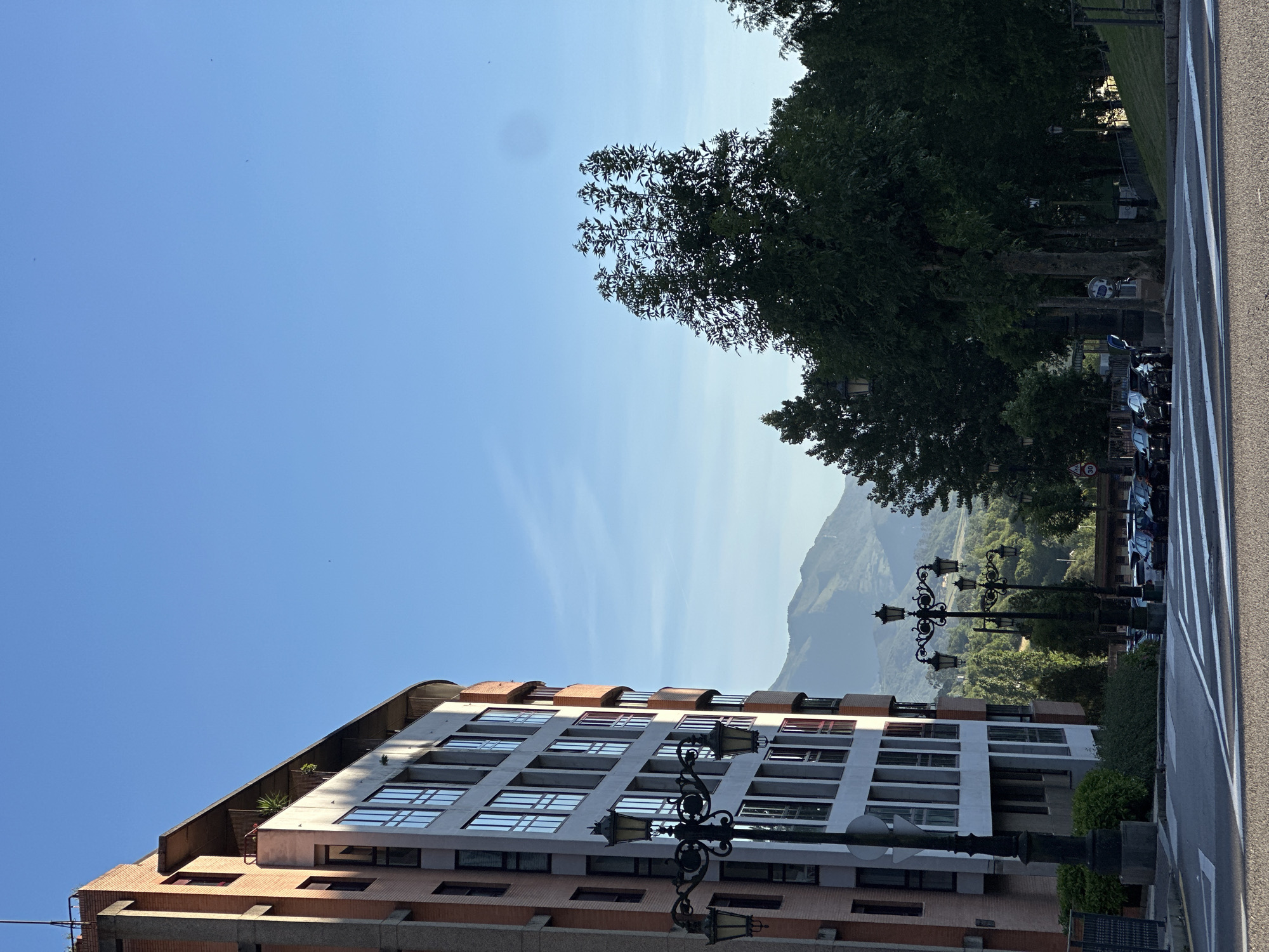The image captures a street scene in Oviedo, Spain, on a clear day. In the foreground, a pedestrian crossing with white stripes is visible on an asphalt road. To the left, a multi-story building with a brick and concrete facade stands tall. The building features numerous windows, some with red frames. A decorative lamppost with four lights is positioned near the building.
In the middle ground, a street lined with trees and more lampposts extends into the distance. Several cars are parked along the side of the street. Beyond the trees and street, a lush green landscape with rolling hills and mountains is visible under a bright blue sky. The mountains in the background are bathed in sunlight, creating a serene backdrop to the urban setting. The overall atmosphere is tranquil and picturesque, suggesting a peaceful morning or afternoon in Oviedo.