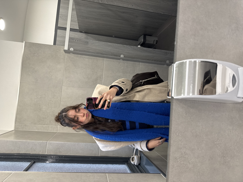 A woman is seen in a mirror selfie, wearing a tan coat, a striped shirt, and a vibrant blue scarf. She holds a phone with a pink case. A black purse sits on top of the coat. Headphones are visible near the bottom edge of the mirror. The reflection shows a bathroom setting with gray tiled walls and metal fixtures. The scene is brightly lit by an overhead light.