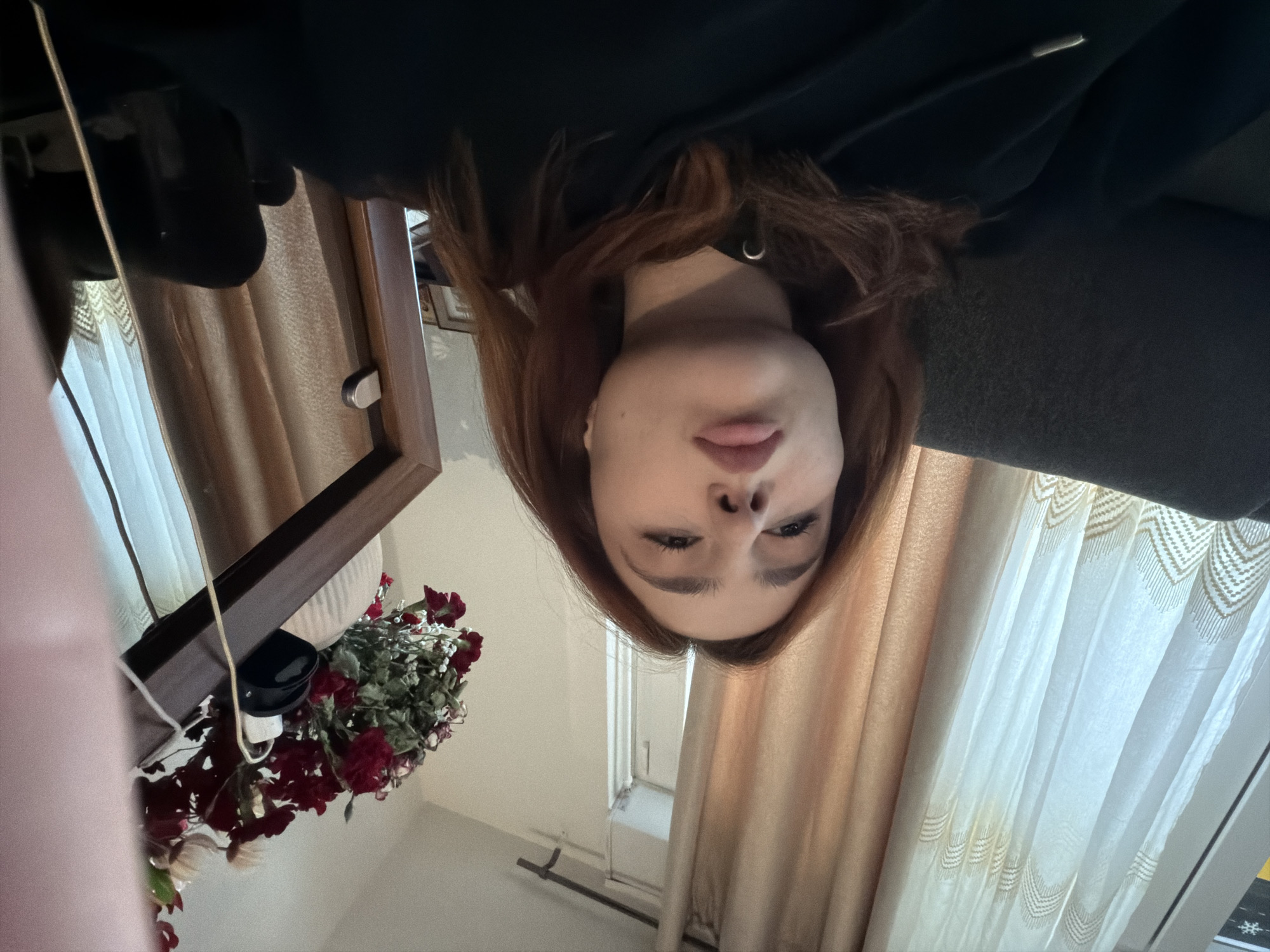 A selfie is taken by a person with red hair, wearing a dark-colored hooded sweatshirt. The camera is positioned at a close-up angle, capturing the individual's face in detail. The person's features include pronounced eyebrows, closed lips, and a visible nose. The backdrop reveals a residential interior with light-colored curtains hanging from a window. A bouquet of red roses is visible in the lower left corner of the frame, positioned near a wooden-framed mirror. The setting suggests an indoor location in Strasbourg, France.