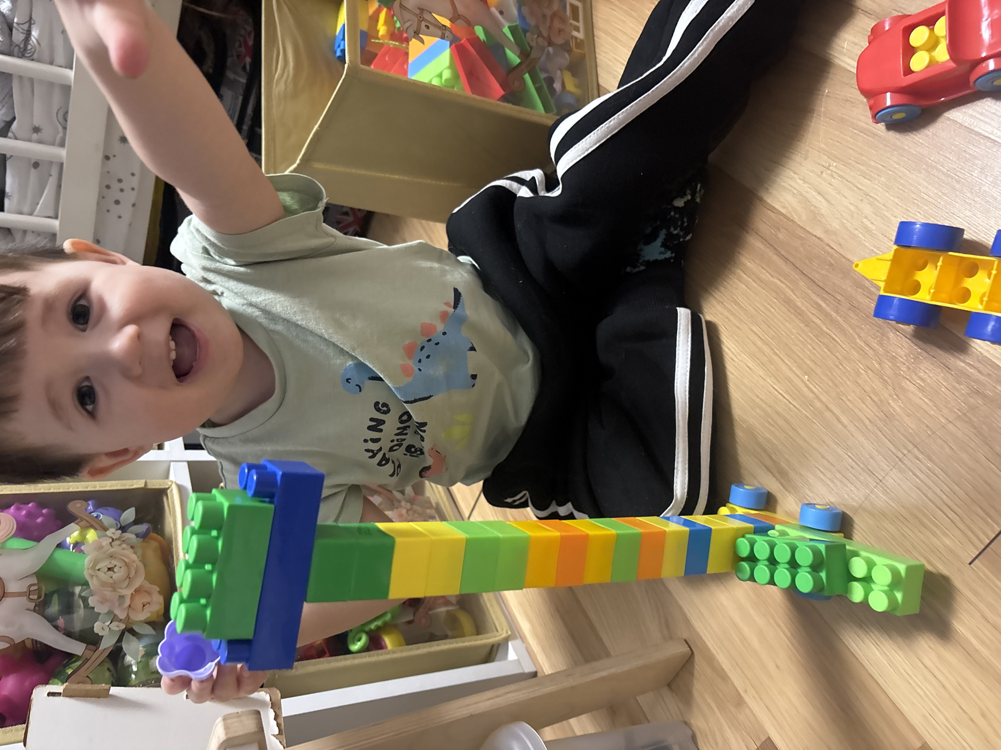 A young boy is captured mid-play indoors on a wooden floor in Chaikovsky, Russia. He is lying on his side, looking directly at the camera with a wide, open-mouthed smile, conveying a sense of joy and engagement. His right arm is raised, holding a long, colorful construction made from large building blocks, featuring alternating green, blue, yellow, and orange pieces.
The boy has light brown hair and brown eyes, and he is wearing a light green t-shirt with a blue dinosaur graphic and the text "KING DINO". He is also dressed in black athletic pants with distinctive white stripes down the sides.
Surrounding him on the light wooden floor are several toys. To his right, two toy vehicles are visible: a red car with yellow accents and a yellow car with blue wheels. Behind and to his left, a light brown fabric bin is filled with an assortment of colorful building blocks. Another partially visible bin on the far left appears to hold more toys, including a rocking horse and some decorative elements. A white crib or playpen railing is also visible along the left edge of the frame. The lighting suggests daytime, illuminating a casual and happy playtime scene.