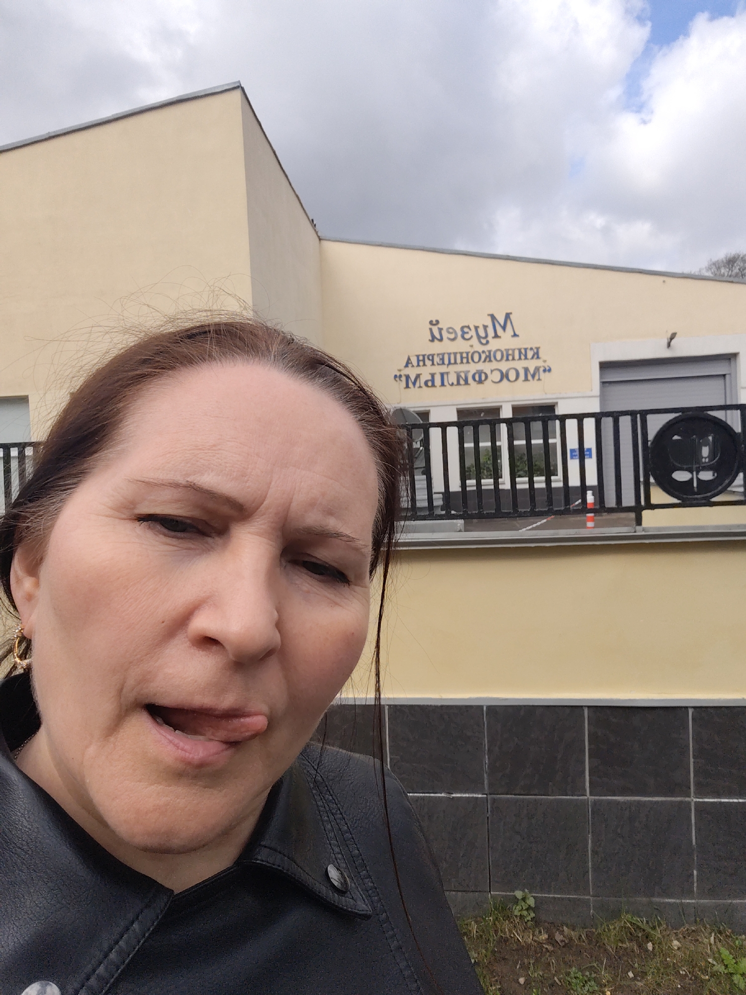 A woman takes a selfie outdoors in front of a building, captured with a playful or silly expression, her tongue slightly extended. She has dark brown hair and is wearing a black jacket, with a golden hoop earring visible on her right ear. Her face shows some lines on her forehead and around her eyes, consistent with her facial expression.
The background features a light yellow or beige building under an overcast sky. Prominently displayed on the building's facade is dark blue text that appears reversed in the image. When mirrored, the text reads: "Музей Киноконцертный "Мосфильм"" (Museum Cinema-Concert "Mosfilm"). This signage identifies the location as the Mosfilm Cinema Concern in Moscow, Russia.
A black metal fence runs along the front of the building, featuring a distinctive circular design element. A closed grey roller shutter is also visible on the building. The immediate foreground shows a dark tiled base leading to a patch of sparse grass and dirt. The scene depicts a cloudy daytime setting.