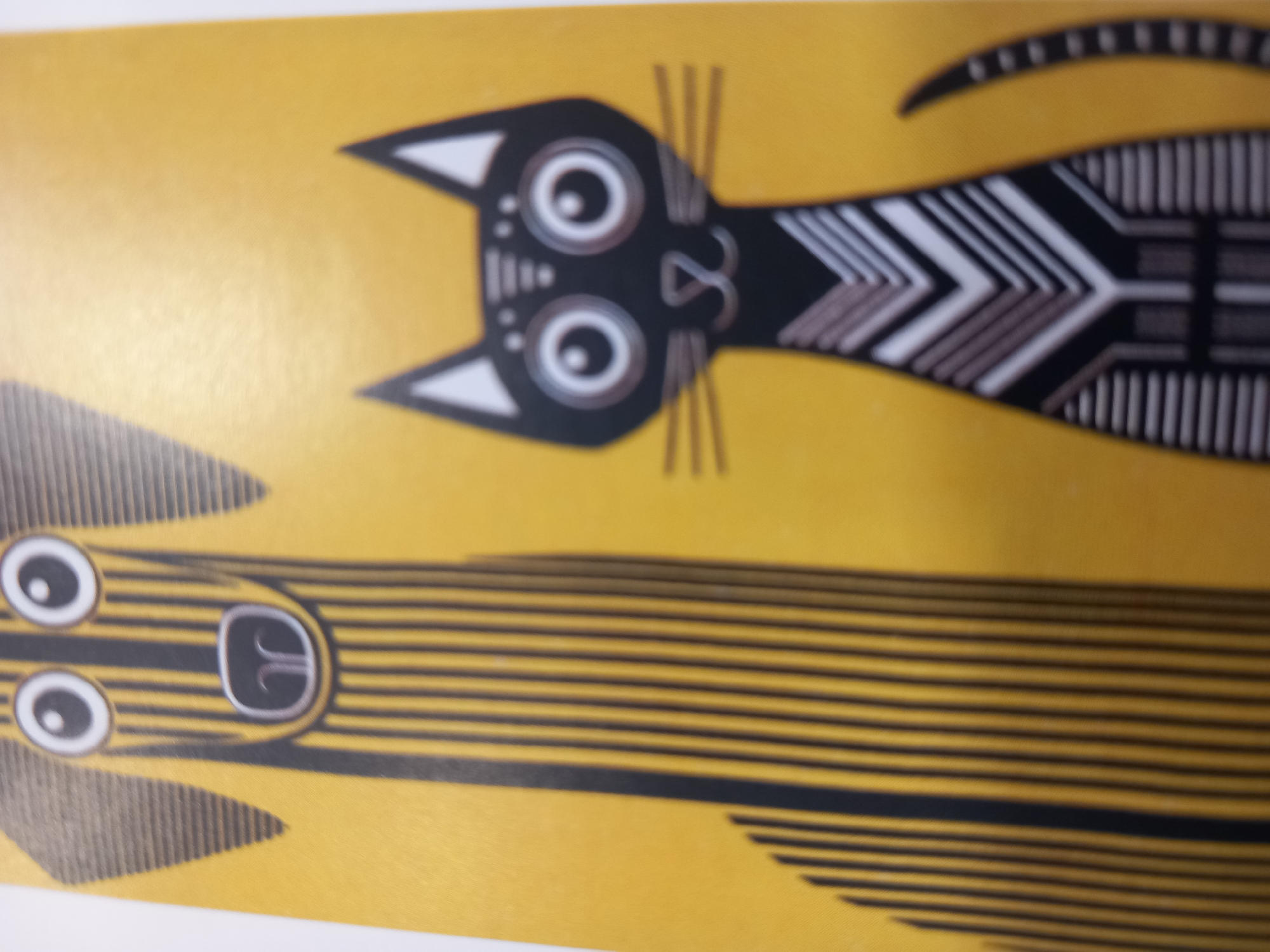 The image is an overhead shot of a yellow background with two stylized black and white cartoon creatures. The top creature appears to be a cat with large, round eyes, pointed ears, and a black body with white and gray striped accents along its back and tail. Its eyes are large and circular with black pupils and white rings, giving it a wide-eyed expression. The cat's face is detailed with small lines suggesting whiskers and a simple mouth. The bottom creature is long and striped, resembling a worm or a snake. It has two large, round eyes similar to the cat's, and a mouth area that looks like a black circle with a white 'E' shape inside. This creature is covered in horizontal black and yellow stripes, with some gray and white zigzag patterns along its body. The style is flat and graphic, with bold outlines and geometric patterns. The overall impression is playful and whimsical, reminiscent of children's book illustrations or abstract art. There is no visible text in the image. The lighting suggests a well-lit indoor environment, but the specific location is unknown.