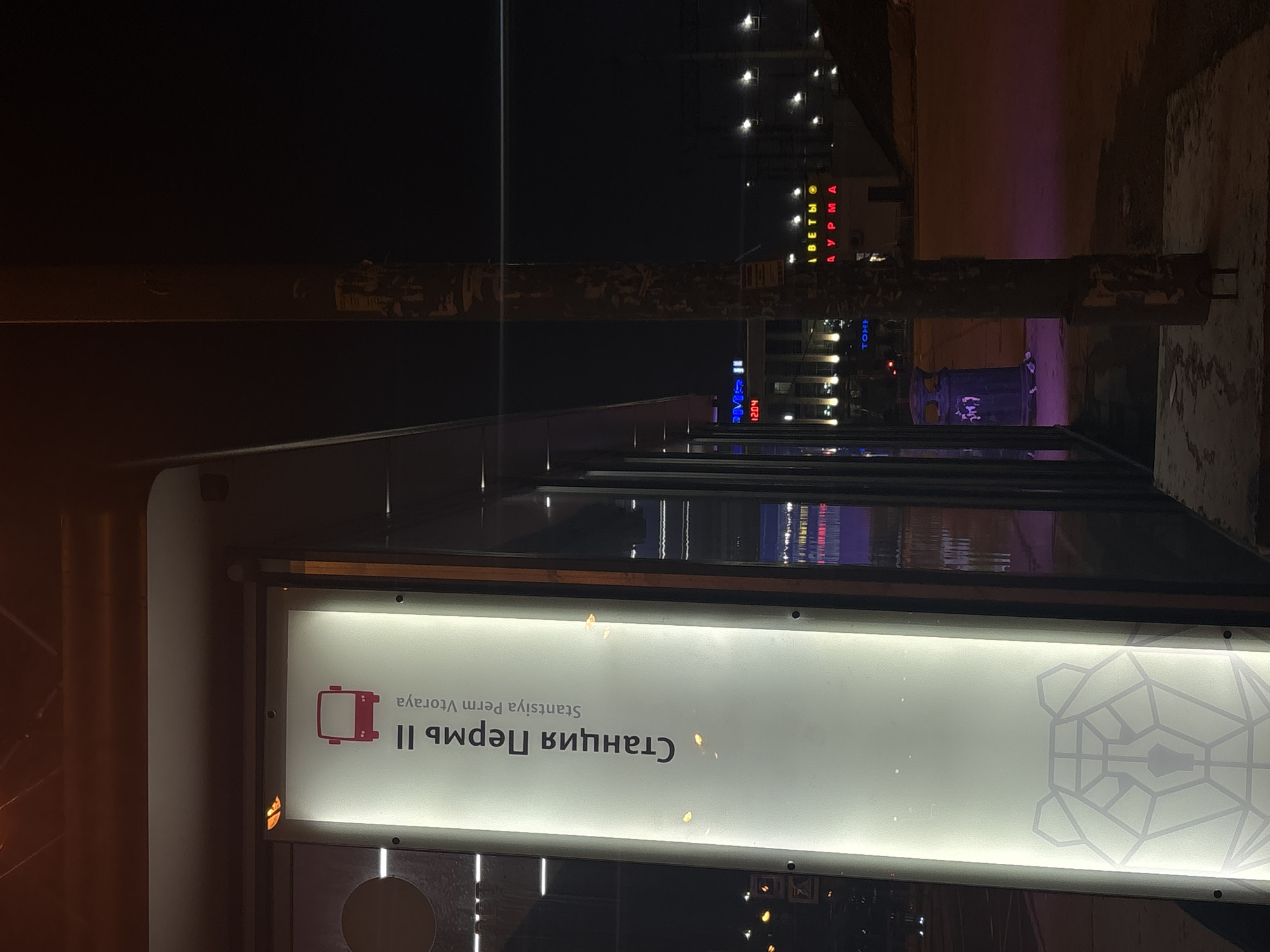 At night, a lit bus stop sign reads "Станция Пермь II Stantsiya Perm Vtoraya," which translates to "Perm II Station" in Perm, Russia. The sign includes a stylized image of a bear head, the symbol of Perm. Beyond the bus stop are illuminated buildings and signs in the distance, suggesting an urban environment.