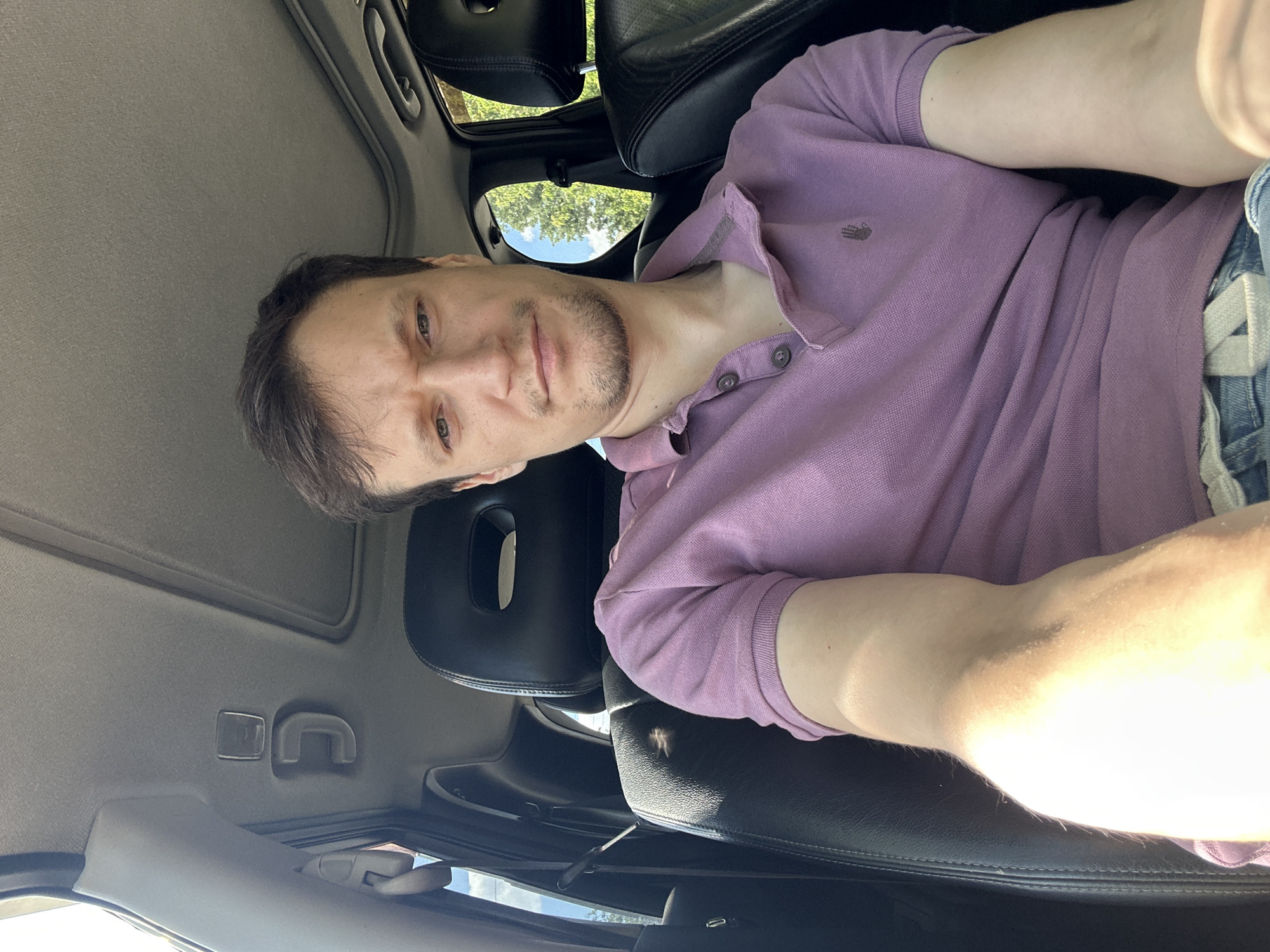 A man is sitting in the driver's seat of a car, taking a selfie. He is looking directly at the camera with a neutral expression. The car's interior is visible, including the headrest of the passenger seat and part of the sunroof. The man is wearing a purple polo shirt and blue jeans. The background outside the car shows trees and a blue sky. There is no indication of the location being in Makiv, Ukraine.