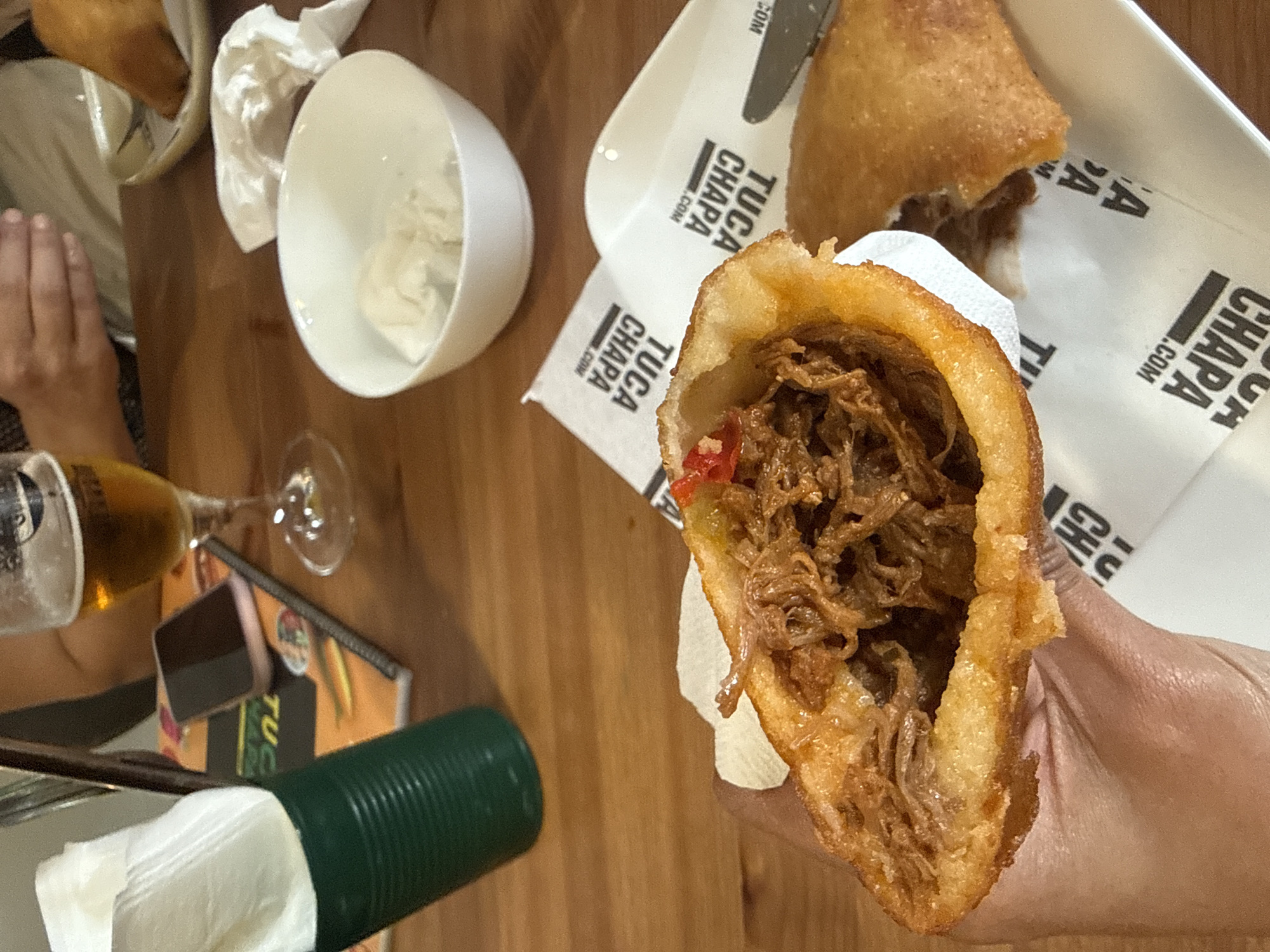 The image depicts a hand holding a halved, fried empanada filled with pulled meat, possibly beef or pork. The empanada rests on a white napkin with the repetition of the logo "TUCA CHAPA .COM". The empanada and its napkin are on top of a white plate that also holds a similar, but complete empanada, part of which has been cut and separated using a metal knife. A small portion of a person's left hand is visible in the lower left, near a glass of beer and a green container, likely a napkin dispenser. A small container with a white substance is positioned above and to the left of the food. The setting is a restaurant in Barcelona, Spain, as evidenced by the logo that is also displayed on a menu near the bottom left. The table is wooden and the scene suggests a casual dining experience.