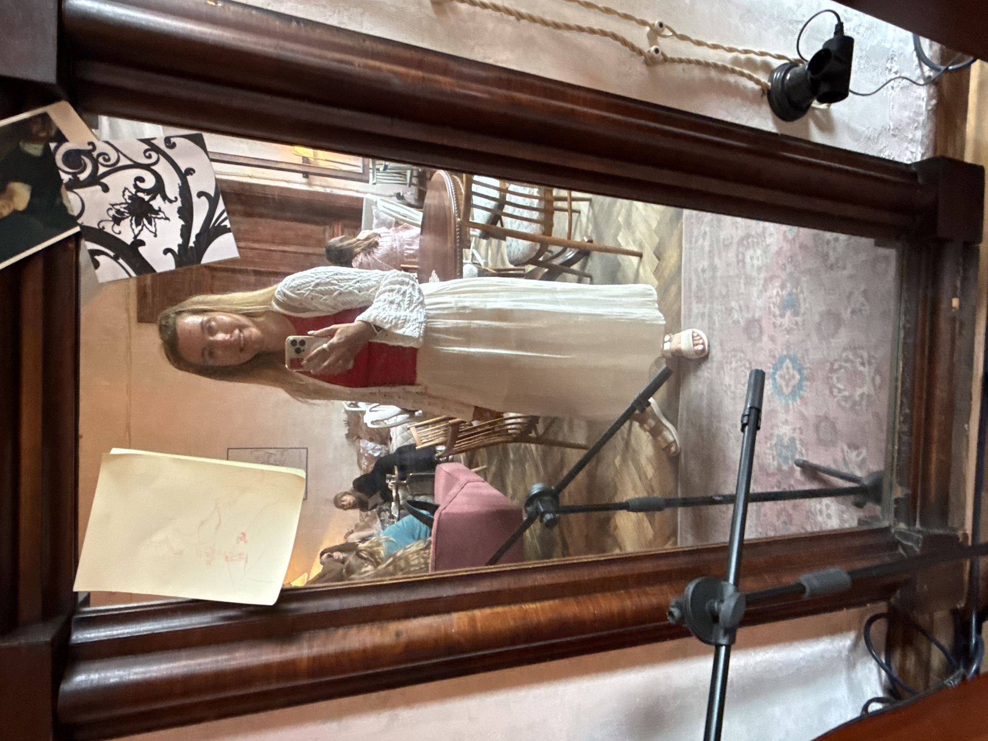A woman stands in front of a large, ornate mirror, taking a selfie with her phone. She is wearing a red top, a white crocheted cardigan, and a long, flowing white skirt. Her hair is blonde and falls to her shoulders. In the mirror's reflection, a room with wooden furniture, chairs, and tables is visible. Several other people are present in the background, seated at tables and on a pink sofa. A pink floral rug covers the floor. The room appears to be a cafe or restaurant, with soft lighting and a warm, inviting atmosphere. The mirror itself is framed by dark wood, with decorative carvings. A microphone stand is positioned in front of the mirror, partially obscuring the lower part of the reflection. There are also pieces of paper and artwork attached to the mirror's frame.