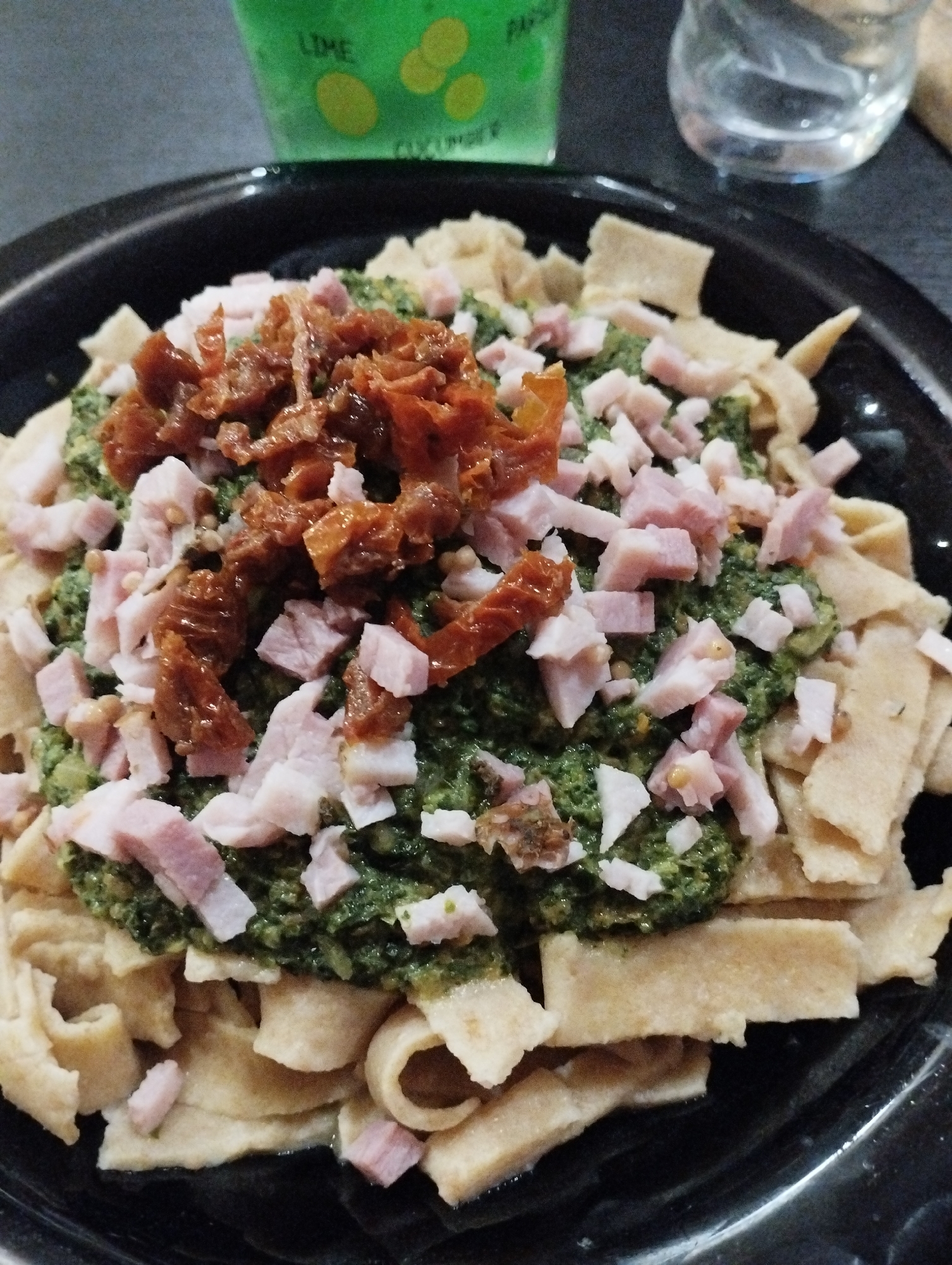 A close-up shot captures a plate of pasta, topped with green pesto, diced ham, and sun-dried tomatoes. The pasta appears to be a wide, flat noodle. A green-labeled beverage is visible in the upper left corner, while a glass of water sits to the right. The scene implies a dining experience, though specific details of the restaurant or setting are not evident. There is no indication about this image being in Burgas, Bulgaria.