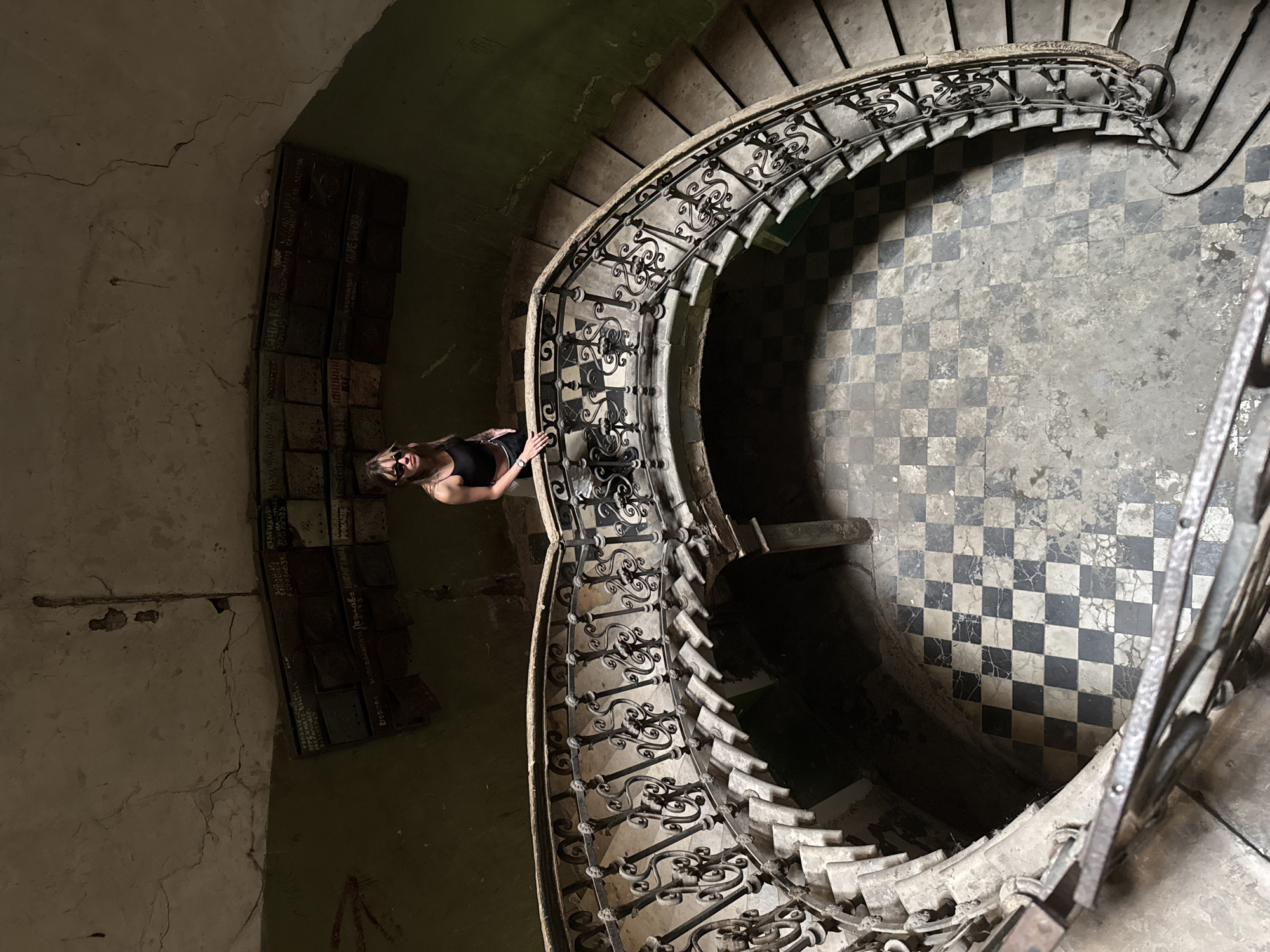An overhead view captures a woman standing on the landing of a grand, decaying spiral staircase in an abandoned building. The woman, dressed in a black tank top and sunglasses, looks down towards the viewer. The staircase itself is ornate with intricate wrought-iron railings and weathered stone steps. The floor below is covered in a checkered tile pattern of black and white squares, also showing signs of age and neglect. The walls are painted a muted green and are cracked and peeling, with rows of what appear to be old mailboxes or plaques affixed to the upper section. This structure is identified as the former House of Journalists in Tbilisi, Georgia.
