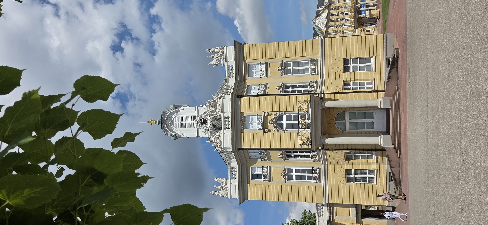 A vertical, medium shot shows the facade of a yellow neoclassical building with ornate white detailing, featuring a central tower with a clock, under a partially cloudy blue sky. Green leaves from a tree hang into the upper left of the frame, obscuring part of the sky and building. The building has multiple stories with regularly spaced windows, many adorned with decorative white elements. A balcony with a golden railing is visible on the second story. Two people are walking on a paved area in front of the building. The person on the left is wearing white pants and a light blue top, carrying a backpack. The person on the right is wearing a khaki shirt and shorts, and a cap. The building appears to be part of the Karlsruhe Palace complex in Karlsruhe, Germany. The ground in front of the building is paved with asphalt, and there are stone steps leading up to the main entrance. A portion of another building with similar architectural style is visible to the right of the main building.