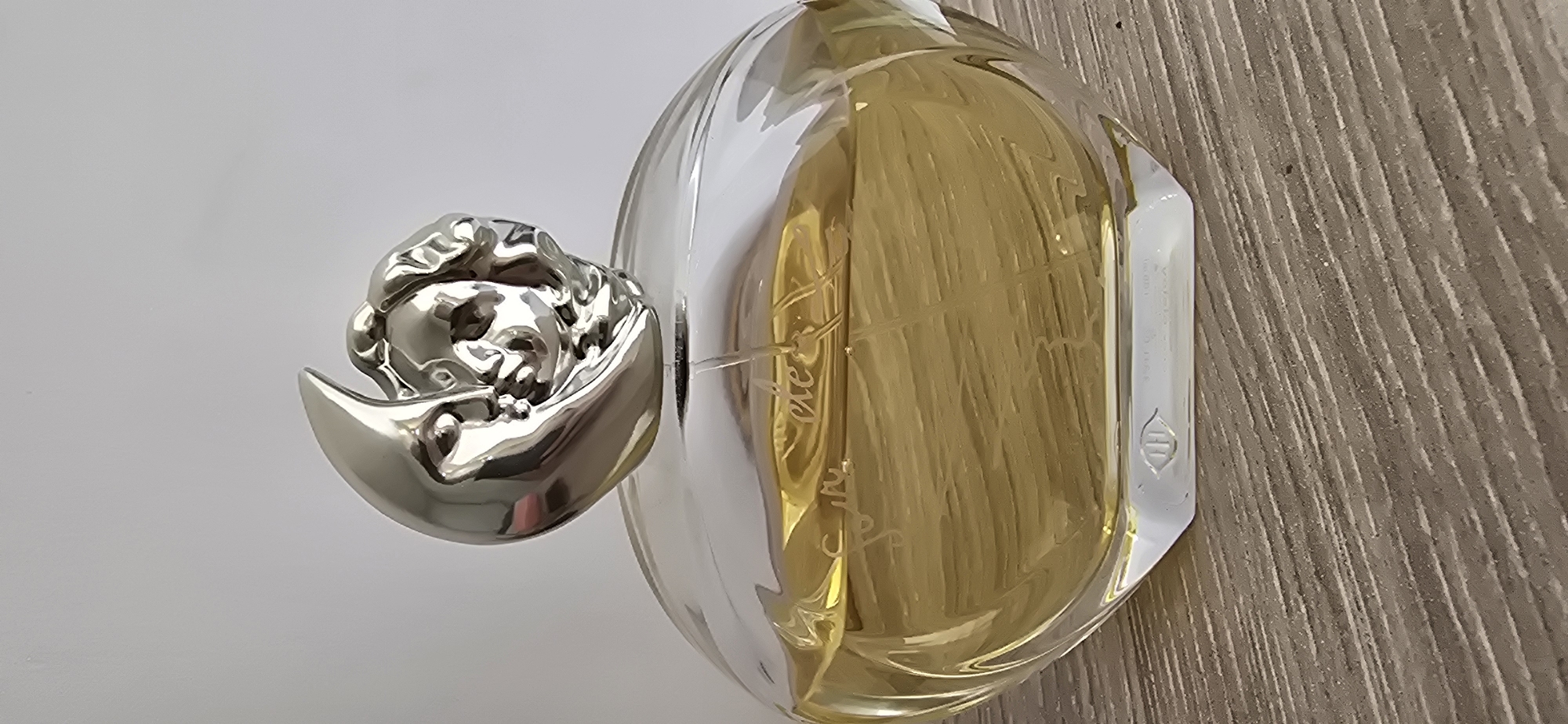 A close-up, eye-level, indoor, waist-up shot focuses on a clear, oval-shaped glass perfume bottle with a metallic, moon-shaped stopper that features a sculpted face. The perfume inside is a light yellow liquid. The bottle is positioned on a wooden surface with a visible grain. The background is a plain, light gray wall. The bottle's glass is etched with a cursive script and a small symbol at the base. The lighting is even and illuminates the bottle from the front. No people are present in the image. The location is not specified within the image itself.