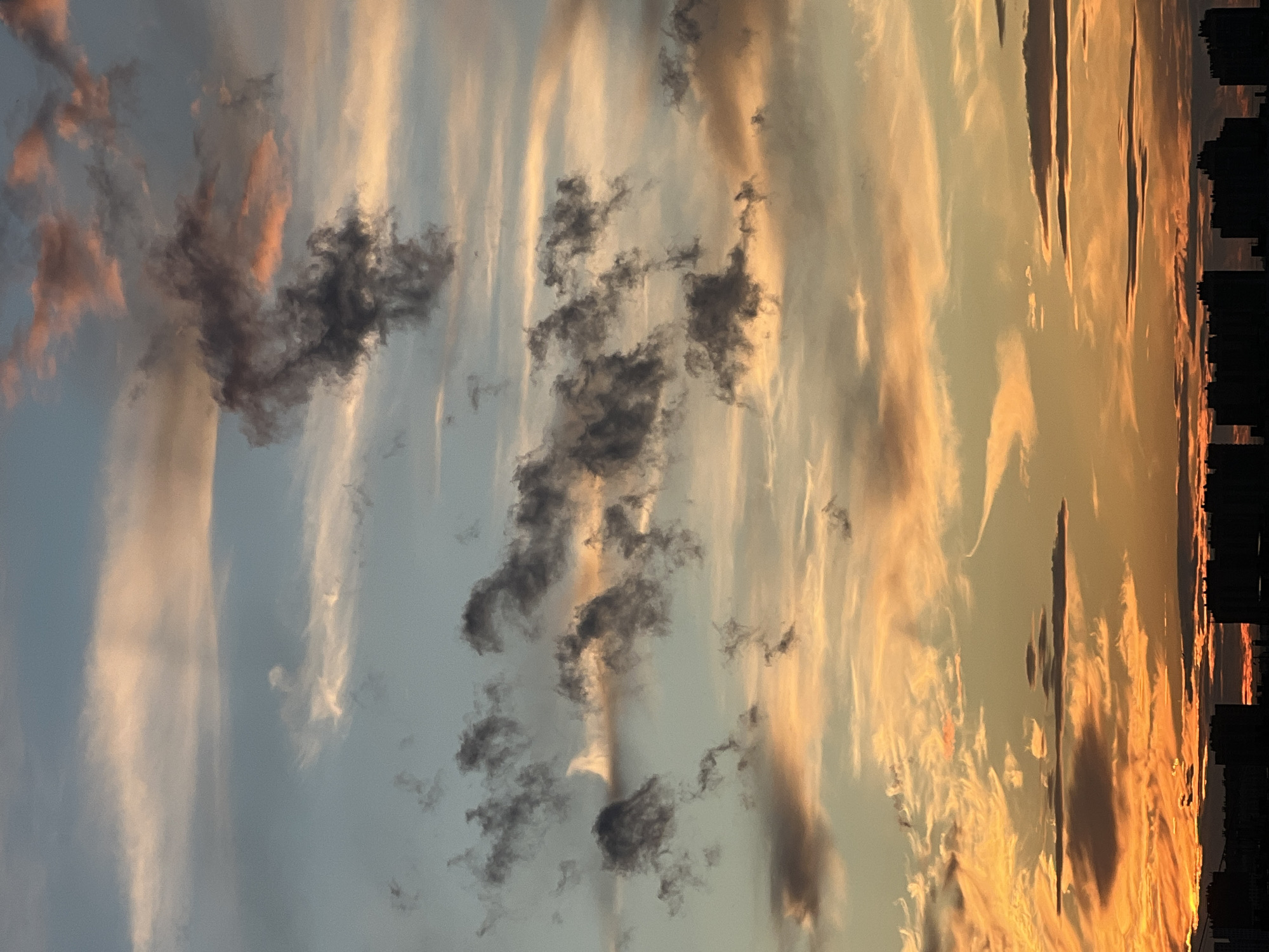 This is a vertical, eye-level, medium shot of a dramatic sunset over a city skyline. The sky is filled with streaks of clouds, with patches of dark, wispy clouds interspersed with brighter, wispy clouds that are lit by the setting sun. The colors of the sky range from a pale blue on the left to a vibrant orange and yellow on the right, indicating it is either sunrise or sunset. On the far right of the image, a silhouetted cityscape is visible, suggesting an urban environment. The clouds appear to be moving, creating a sense of dynamism and awe. The overall mood is one of natural beauty and the power of the elements. There is no visible text in the image.