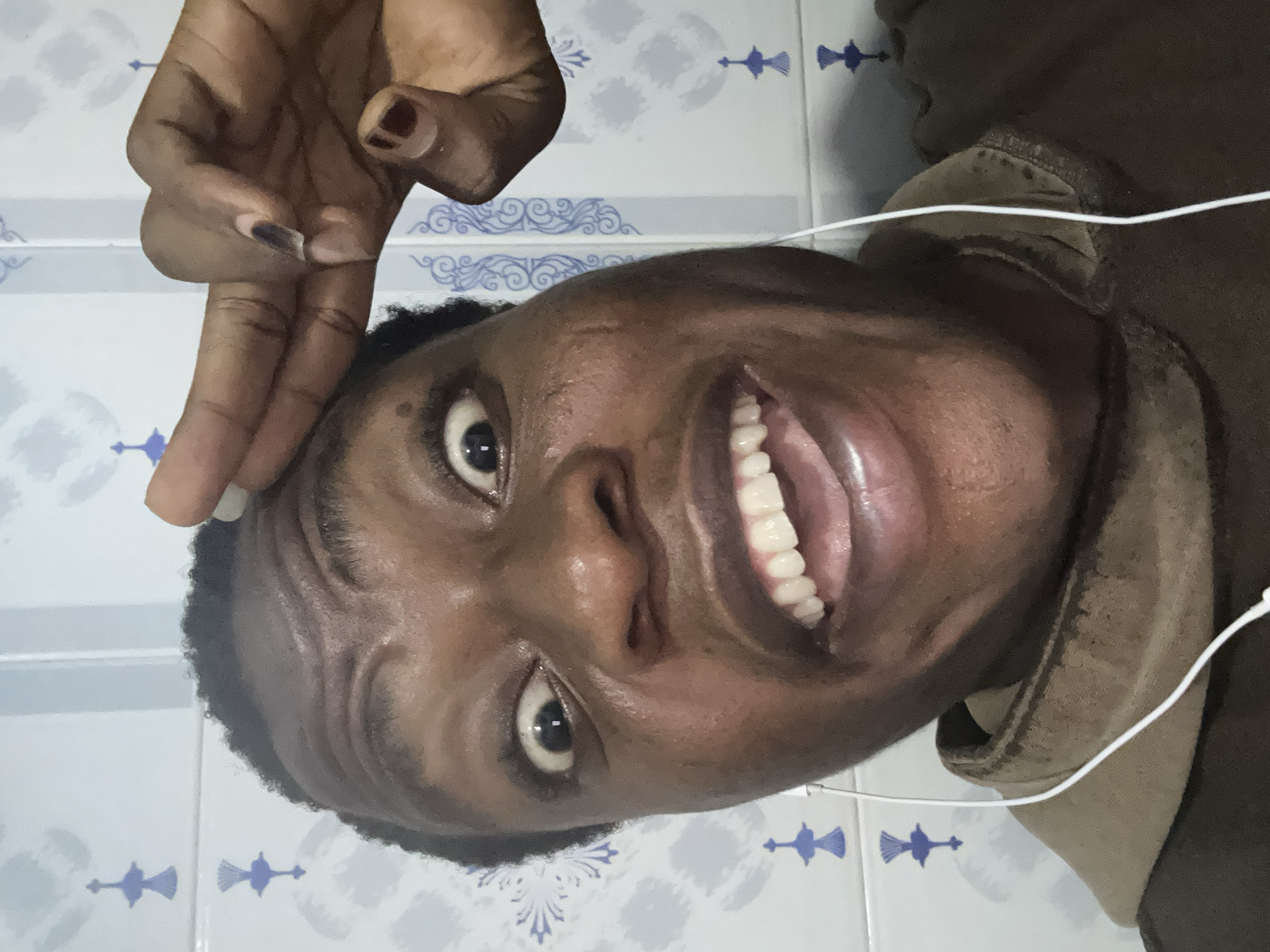 A close-up shot depicts a smiling dark-skinned person with visible teeth and white earbuds. Their hand is positioned above their head. The background features tiled walls with a blue and white pattern. The location is not specified.