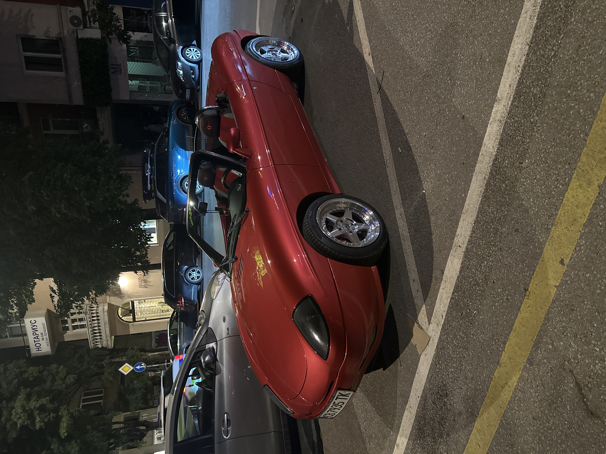 A metallic red Fiat Barchetta convertible is parked on a street at night in Sofia, Bulgaria. The car is in the foreground, angled towards the viewer's left. Its black convertible top is down, revealing two red and black bucket seats. The car has chrome multi-spoke wheels and a license plate that reads "CB 7935 TK".
To the left of the Fiat, a silver sedan is partially visible. Behind the Fiat, several other cars are parked, including a dark-colored station wagon and a blue SUV with a roof rack.
The background features a building with a sign that says "НОТАРИУС" (Notary) in white lettering. Below the notary sign, there is a balcony with a white railing. Adjacent to the notary office, there is a shop with "MI AMOR" signage. The street is paved and marked with white and yellow parking lines. Trees with dark foliage are visible in the background, obscuring parts of the buildings. The scene is lit by streetlights and the headlights of the vehicles.