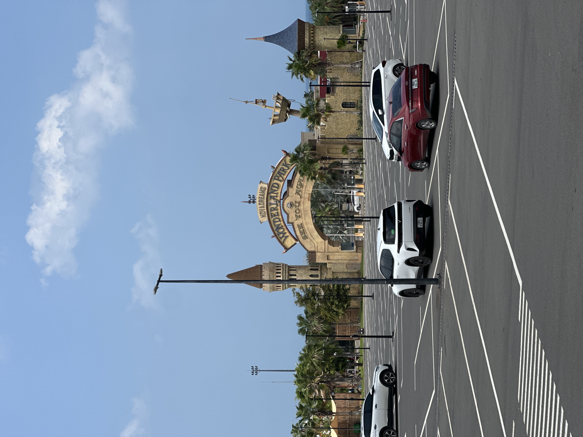 The image captures an expansive, largely empty asphalt parking lot in the foreground, leading to the entrance of an amusement park in the midground, under a clear blue sky with scattered white cumulus clouds.
The central subject is the entrance to "NOVADREAMS WONDERLAND PARK," with "SEE YOU AGAIN" inscribed below the main signage. The entrance features a large archway flanked by two distinct towers constructed from light brown stone-like material. To the left, a tall, cylindrical tower with a conical brown roof stands prominently. To the right, a smaller turret with a steep blue roof is visible. Between the arch and the blue-roofed turret, a large, weathered ship's mast with a crow's nest is dramatically angled, resembling a shipwreck display. Palm trees are visible behind and around the park structures. In the far background, a tall, dark metal tower with floodlights is present to the left of the main park entrance.
The parking lot, marked with numerous white lines delineating parking spaces, is sparsely occupied. In the foreground-left, a white Peugeot 3008 SUV is parked. In the foreground-right, a white VinFast VF 7 electric SUV is parked, displaying the Vietnamese license plate "49A 838.63." Further back on the right, a red Hyundai Elantra sedan is parked, showing the Vietnamese license plate "86A 284.26." Another white vehicle is partially visible on the midground-left. Numerous black lampposts, designed for parking lot illumination, are distributed throughout the scene, with one prominent lamppost centrally located in the foreground. A white zebra crossing pattern is visible on the asphalt in the bottom-left.
No people are visible in the scene. Based on the "NOVADREAMS WONDERLAND PARK" signage and the Vietnamese license plates (49A for Lâm Đồng Province, 86A for Bình Thuận Province), the location is identified as Tien Thanh Ward, Phan Thiet City, Bình Thuận Province, Vietnam, likely associated with the NovaWorld Phan Thiet development.