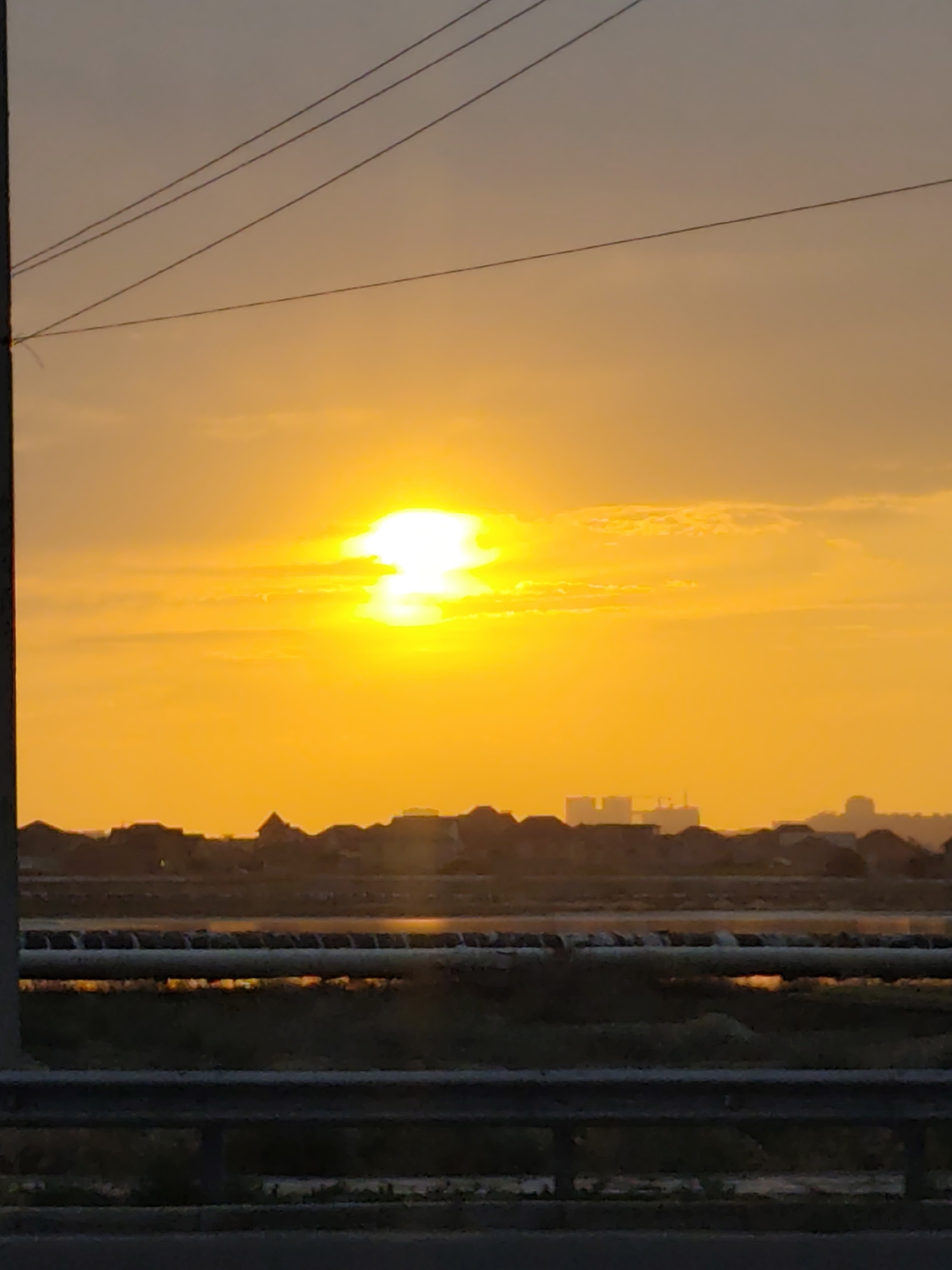 A vertical, full-frame photograph captures a sunset over a semi-urban landscape in Ömirzaq, Kazakhstan. The sun, a bright, almost white orb, sits high in the sky, radiating a powerful orange glow that saturates the atmosphere. Wispy clouds are visible, catching the light and creating a soft, diffused effect. In the foreground, a dark, silhouetted pole with power lines stretches upwards from the left side of the frame, crossing the bright sky. Below the sky, a band of darker, silhouetted structures forms the skyline of a town or city. These appear to be buildings with varied rooflines, suggesting residential or mixed-use development. Further down, a dark, hazy band suggests a body of water or a low-lying area. In the immediate foreground, a dark, metallic barrier or railing runs horizontally across the bottom of the image, partially obscuring the view of the landscape beyond. The overall impression is one of twilight settling over a developing area, with the vibrant sunset providing a dramatic backdrop.