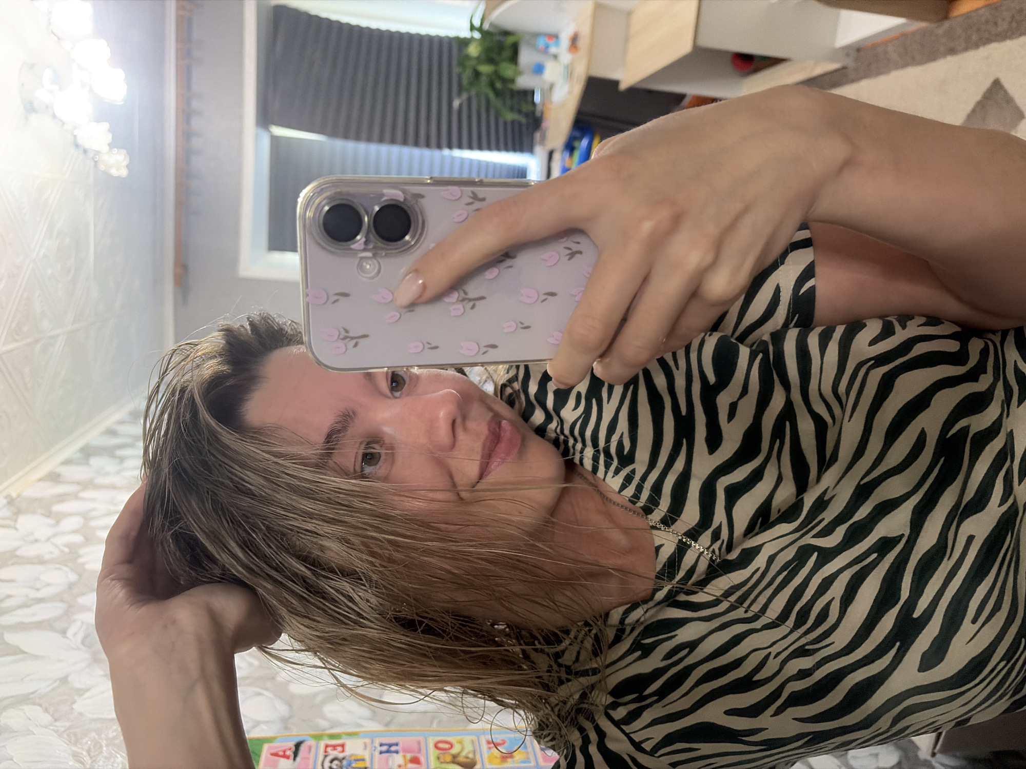 A young woman with wet, blonde hair is taking a selfie in a room with patterned wallpaper and a tiled ceiling. She is wearing a zebra-print t-shirt and a silver necklace. She is holding a phone with a purple floral case in her right hand, and her left hand is in her hair. In the background, a mirror reflects part of the room, including blinds on a window and a shelf with plants and other items. The scene is a residential interior, likely a bathroom or bedroom, given the mirror and vanity area visible in the reflection. There is no identifiable information to place this image in Chaikovsky, Russia.