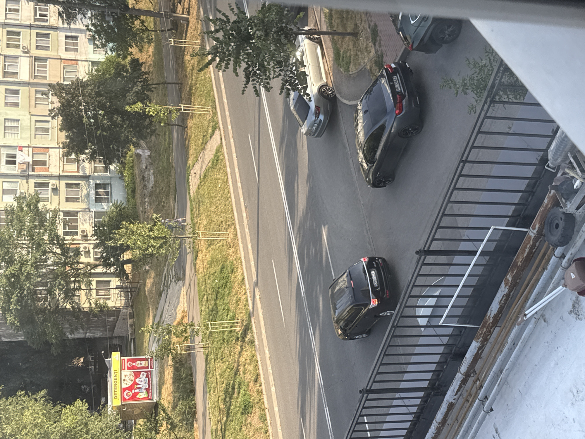 The image captures a street scene in Chișinău, Moldova, likely during daylight hours. A black SUV is parked on the right side of the street, with its rear facing the camera. Ahead of it, a dark gray coupe is parked in what appears to be a turning lane, also with its rear facing the camera. Further up the street, a silver sedan is parked on the right side of the road, angled towards the curb. A black SUV is visible in the distance on the far right, partially obscured.
In the background, a multi-story residential building with light-colored facades and numerous windows dominates the skyline. Trees and grass line the street and the area between the road and the building. A small kiosk with a yellow roof and red signage reading "DETERGENTI" is visible on the left side of the frame, displaying images of cleaning products. A person is walking on the sidewalk in the middle ground on the left.
The foreground shows a metal railing and a rooftop or balcony with pipes and some debris, indicating a raised vantage point for the photographer. The overall environment suggests an urban residential area.