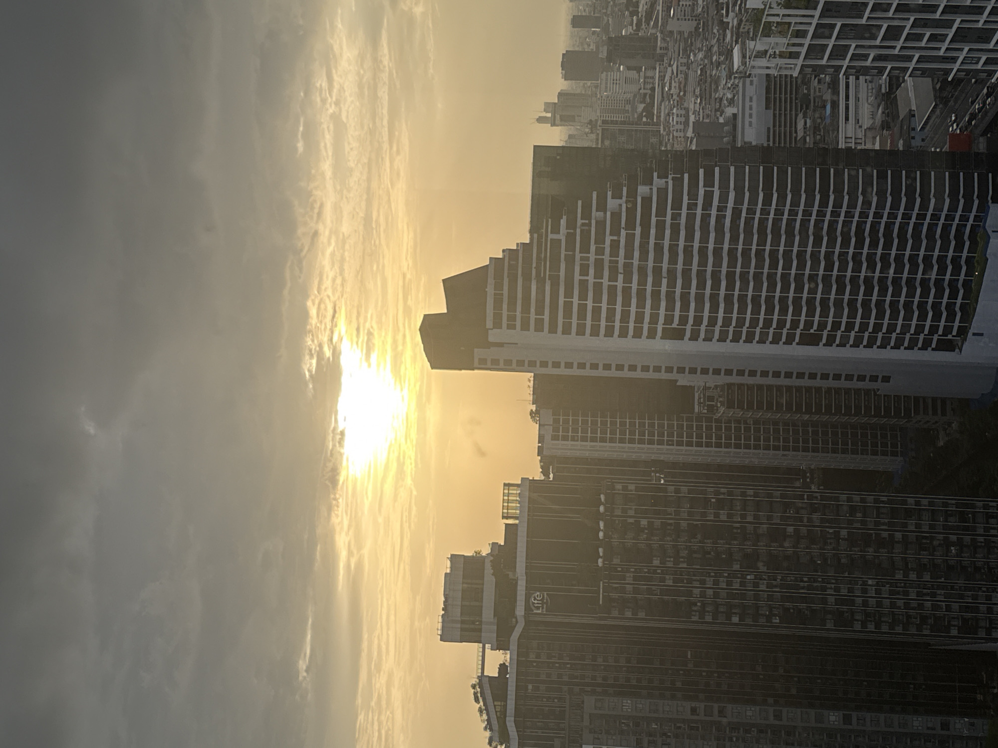 This image captures a view of the Bangkok skyline at sunset. The sun is partially obscured by clouds, casting a warm, golden light across the scene. In the foreground, several modern high-rise buildings dominate the view. One building on the left features a prominent sign with the word "Life" in white letters. Another building on the right has a distinctive tiered or stepped design. In the background, more buildings of varying heights are visible, creating a dense urban landscape. The sky is filled with clouds, some of which are illuminated by the setting sun, creating a dramatic effect. The overall impression is of a bustling metropolitan city at dusk.