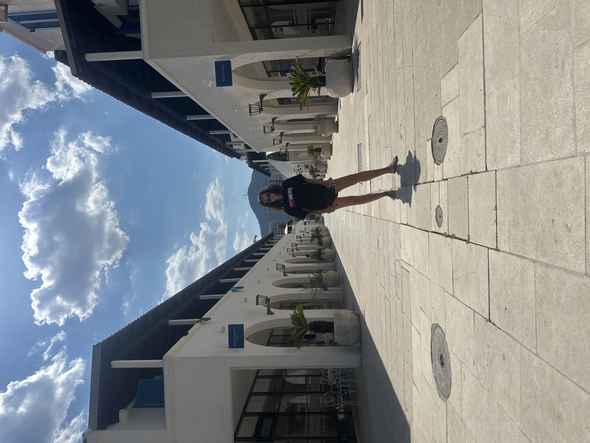 A woman stands in the middle of a pedestrian walkway lined with white buildings. The walkway is paved with light-colored stone tiles and there are circular metal grates on the ground. The buildings on either side of the walkway have arched doorways and windows. There are potted plants placed outside the buildings. In the background, there is a mountain with a television tower on top. The sky is blue with white clouds. The woman is wearing a black t-shirt, dark shorts, and sandals. She is facing the camera and has sunglasses on. The overall scene suggests a sunny day in a resort town. This location appears to be the promenade of the Dukley Gardens complex in Budva, Montenegro, known for its luxury apartments and waterfront access.
