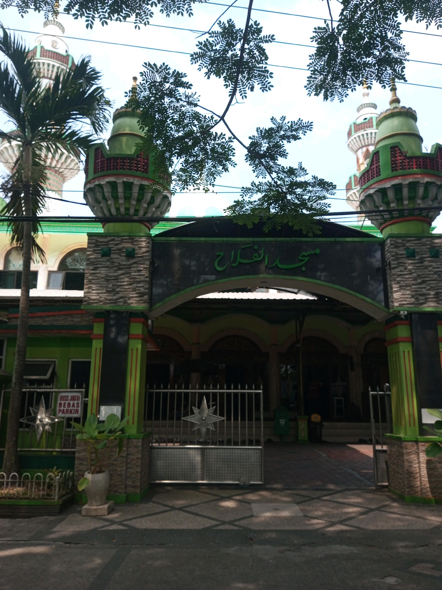 The image shows the entrance to the Al-Falah mosque, located in Malang, Indonesia. The mosque features a prominent green and gray facade with ornate minarets. Arabic script, likely indicating the mosque's name, is visible above the entrance archway. A metal gate with a star-shaped design is open, revealing the pathway leading into the mosque. Trees and overhead wires are present in the foreground, partially obscuring the view of the minarets. A sign that reads "KHUSUS JAMAAH BEBAS PARKIR" (meaning "For Congregation, Free Parking") is affixed to a post near the entrance. The scene appears to be a street-level view on a sunny day.