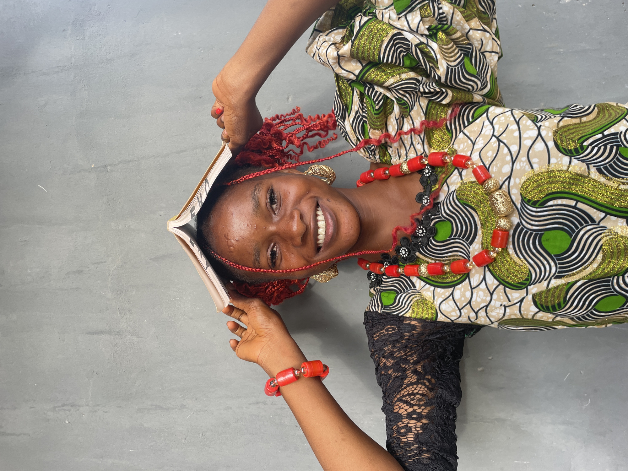 A vibrant, horizontally oriented portrait captures a young Black woman smiling joyfully at the viewer. She is positioned against a plain, light gray surface, possibly a concrete floor or wall. Her head is tilted, and she holds an open book above her head, partially framing her face. Her hair is styled in intricate red braids or extensions, with some strands flowing loosely.
She is dressed in a colorful, patterned fabric, likely a traditional dress or top, featuring a striking design with green, black, white, and tan/beige colors, accentuated by shimmering or glittery green sections. One visible sleeve is made of black lace. Her attire is complemented by ornate traditional jewelry, including a prominent red beaded necklace with gold and black decorative elements, and a matching red beaded bracelet on her right wrist. A decorative gold earring is also visible on her left ear.
The woman's expression is one of bright happiness and confidence, with a wide, genuine smile revealing her white teeth. The partial word "EKO" is visible on the pages or spine of the book she is holding. The lighting is bright and even, highlighting her features and attire. The image conveys a sense of cultural celebration and joyful spirit, set within the context of Badagry, Nigeria.