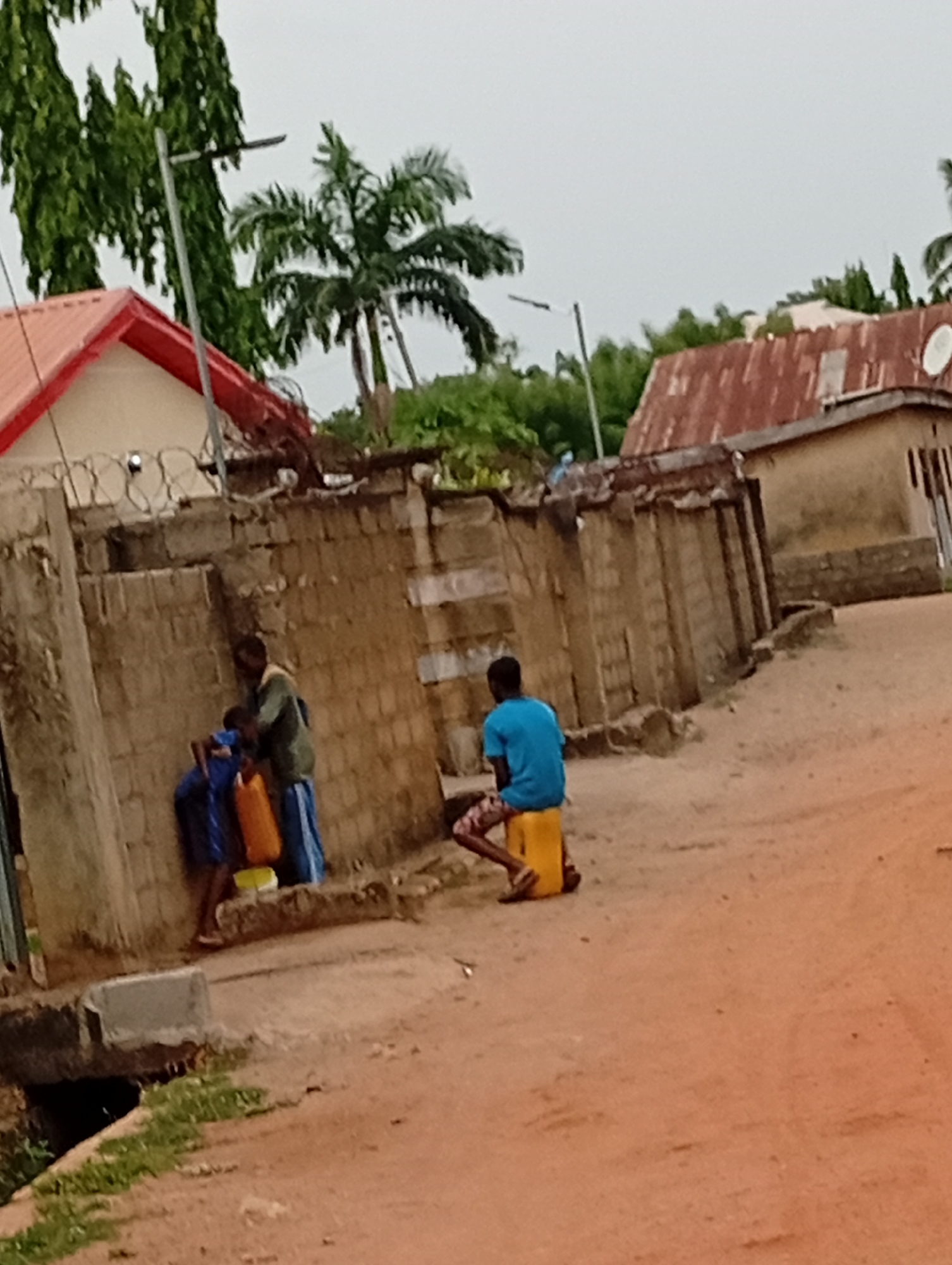 In Adankolo, Nigeria, the scene depicts a dusty, unpaved road lined with dilapidated brick walls and modest buildings. A palm tree is visible in the background. At the left, two individuals stand near a source of water, filling containers. One person wears blue and carries a container. A second individual, wearing an olive-green shirt and a blue skirt, assists. A third individual, wearing a blue t-shirt, sits on a yellow container.